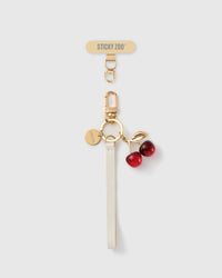 STICKYZOO ivory leather phone strap with a glossy red cherry charm, gold-tone leaf, engraved disc and a gold Phone Strap Card connector tab.