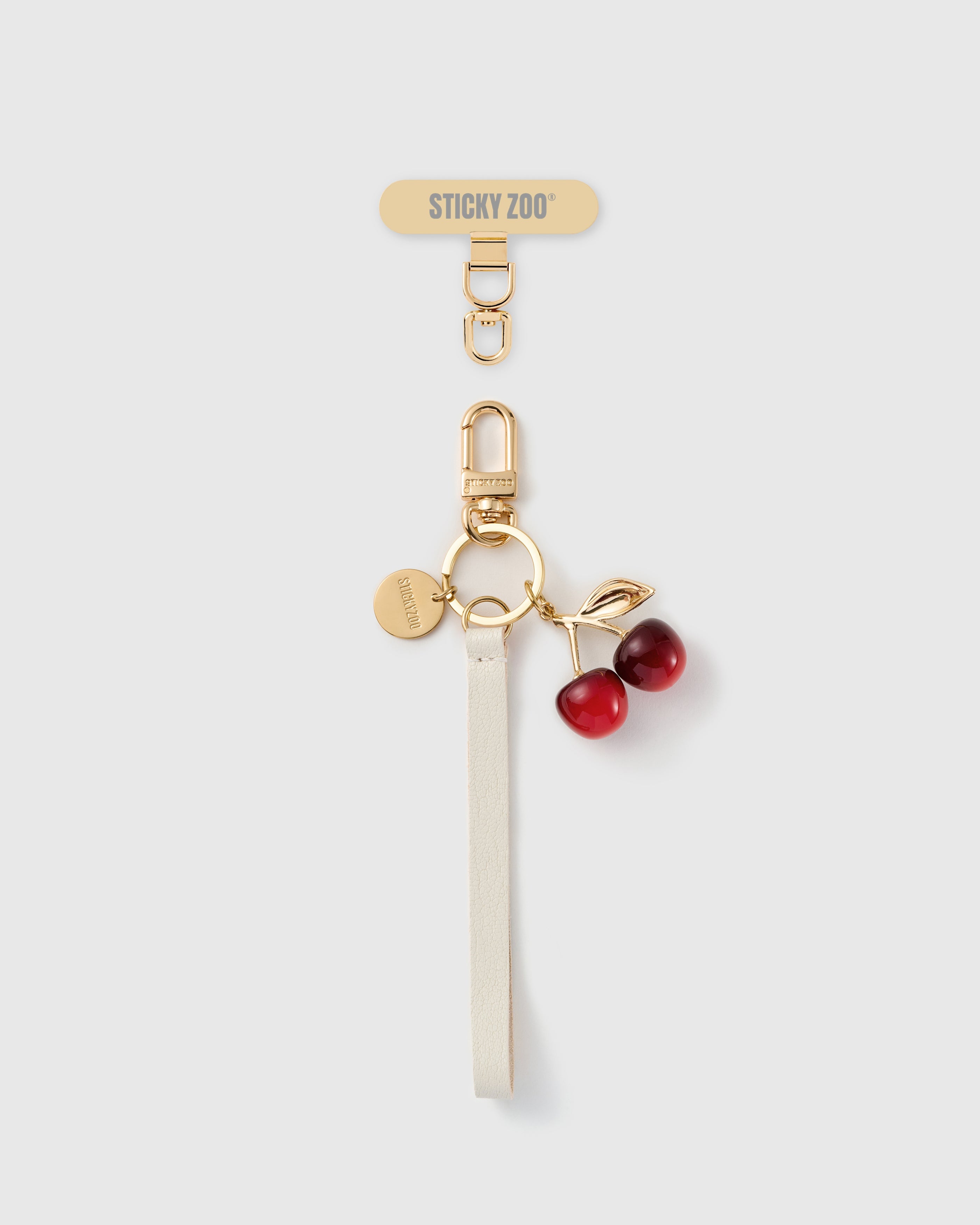 STICKYZOO ivory leather phone strap with a glossy red cherry charm, gold-tone leaf, engraved disc and a gold Phone Strap Card connector tab.