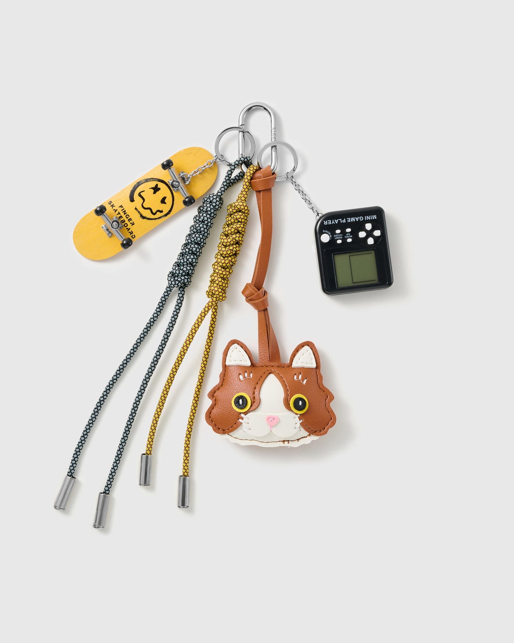 Tuxedo Cat Skate Stack Charm with a tuxedo-cat face, skateboard, black handheld-game motif and grey-and-yellow cords on a light background