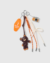 New York Teddy Camera Stack Charm with a brown teddy, orange new york tag, miniature camera and orange-and-cream cords on a light background