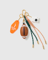 New York Football Stack Charm with an american-football motif, orange new york tag, miniature camera and green-and-orange cords on a light background