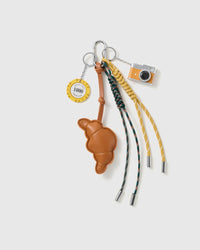 Croissant Lucky Camera Stack Charm with a croissant, lucky chip motif, miniature camera and green-and-yellow cords on a light background