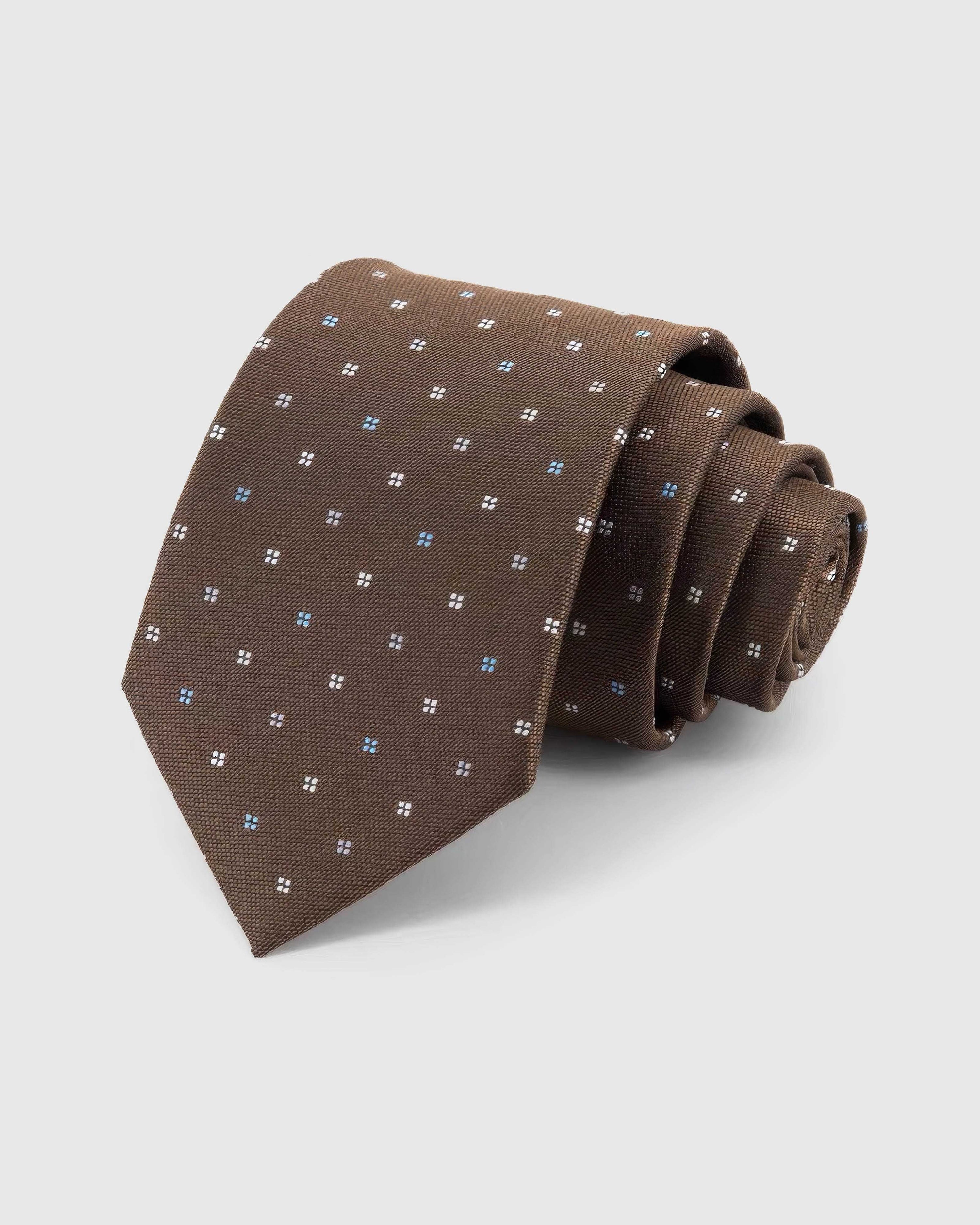 STICKYZOO Walnut Quatrefoil Tie rolled on white, showing small ivory and pale-blue quatrefoils across 100% mulberry silk in walnut brown