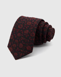 STICKYZOO Noir Crimson Bloom Tie rolled on white, showing a dark ground layered with crimson botanical blooms and shadowed leaves across the front blade.