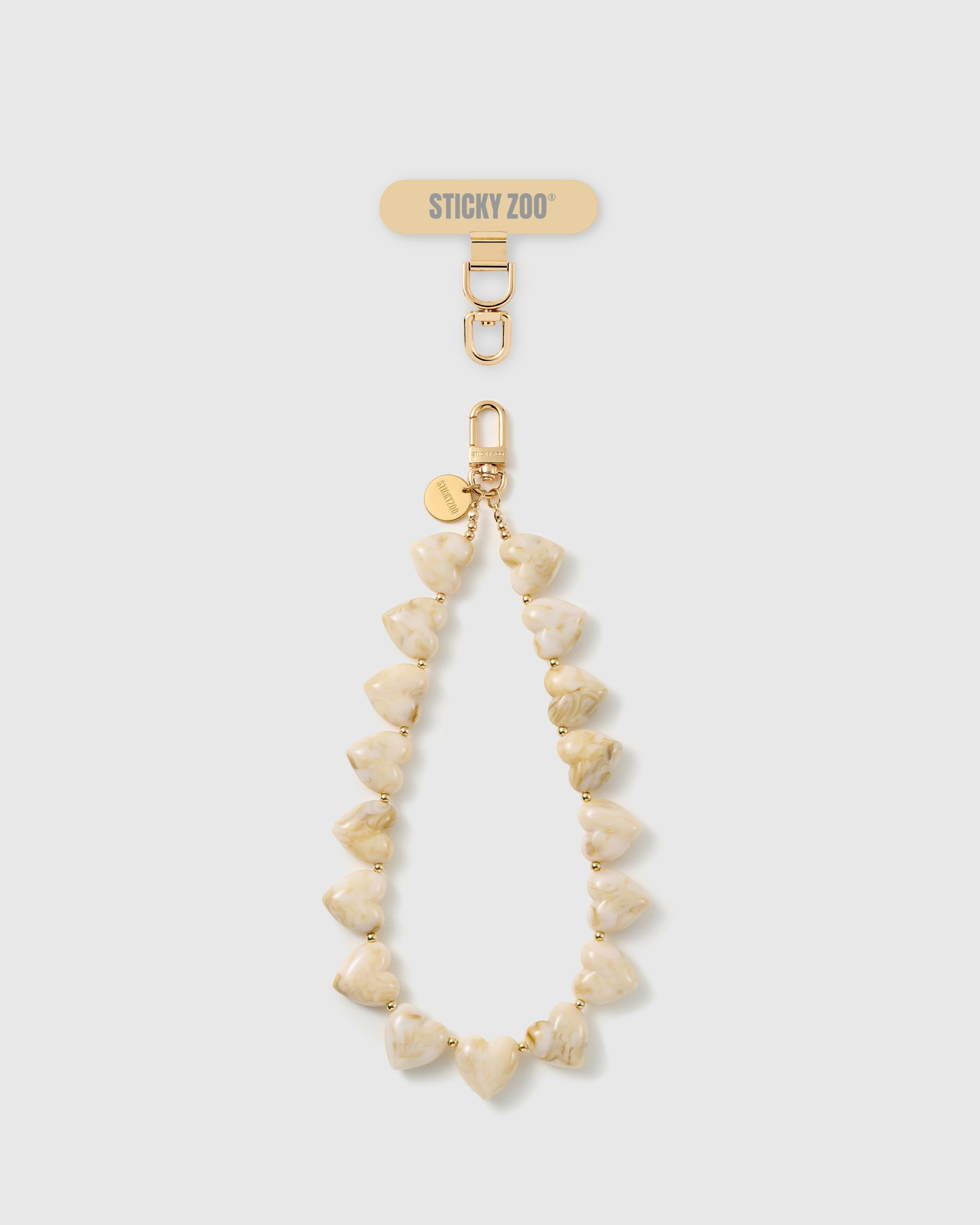 Marbled ivory heart bead phone strap laid straight, with a gold hook, an engraved gold disc, and a STICKYZOO phone-strap card at the top.