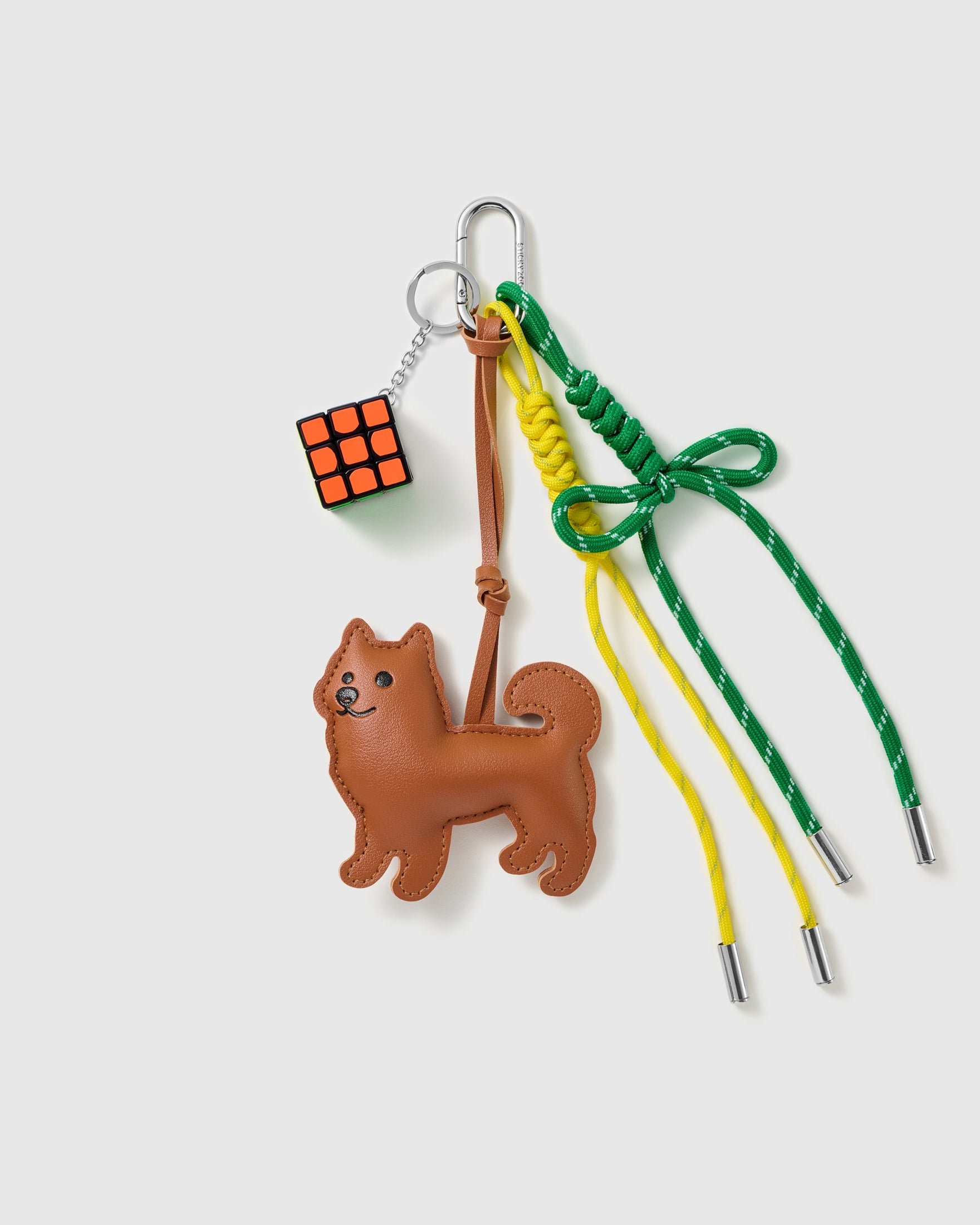 Shiba Puzzle Stack Charm with a brown shiba-style pup, puzzle cube, green bow and yellow-and-green cords on a light background