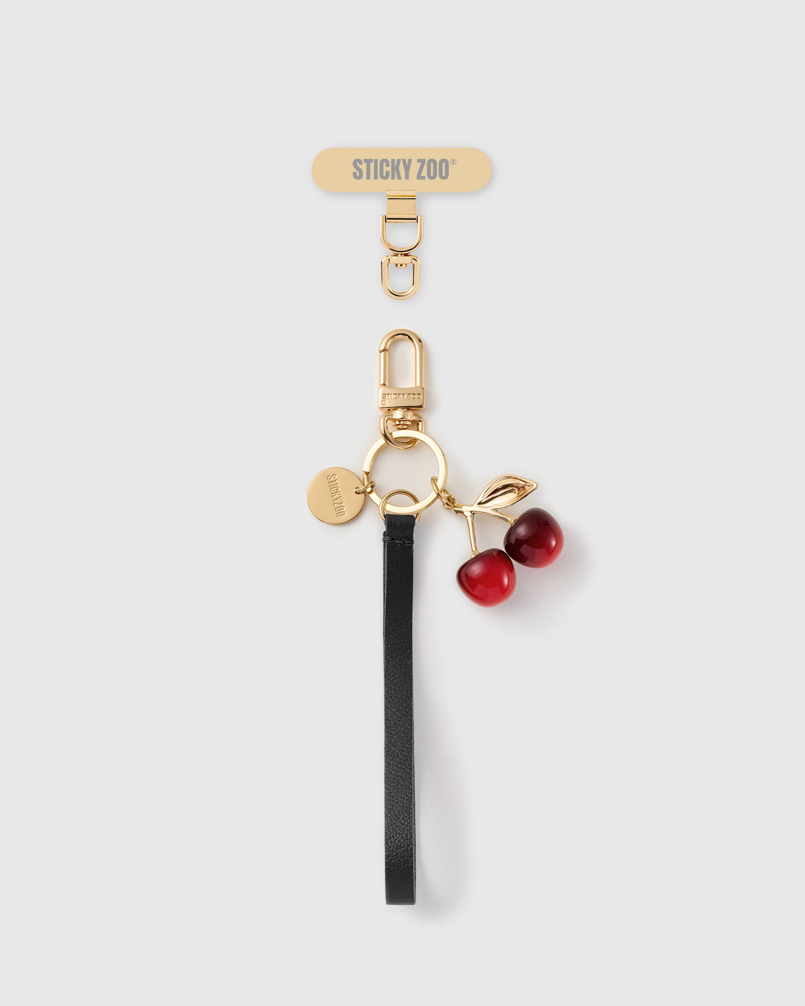 STICKYZOO black leather phone strap with a glossy red cherry charm, gold-tone leaf, engraved disc and gold Phone Strap Card tab.