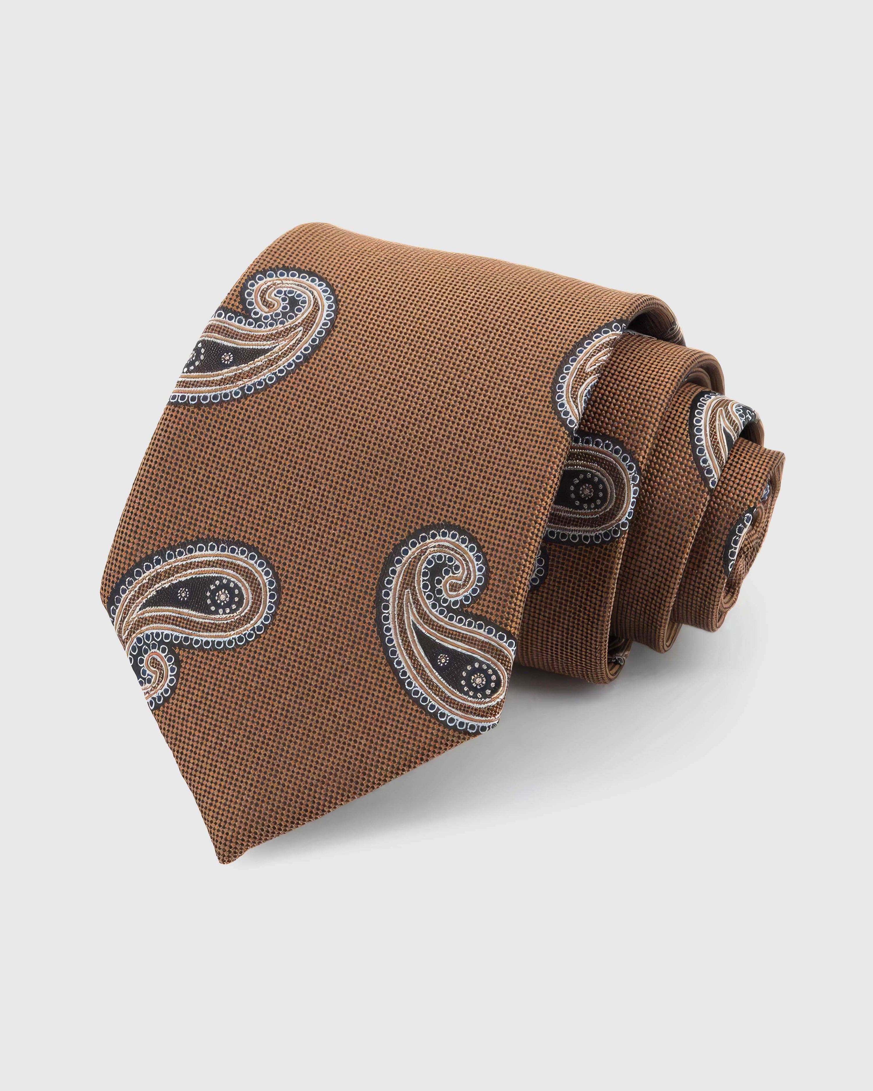 STICKYZOO Cognac Paisley Tie rolled on white, showing a cognac-toned ground with sweeping black, cream, and brown paisley forms across the front blade.