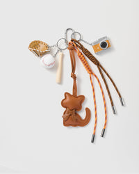 Cocoa Cat Camera Stack Charm with a cocoa-brown cat silhouette, baseball motifs, miniature camera and orange-and-brown cords on a light background
