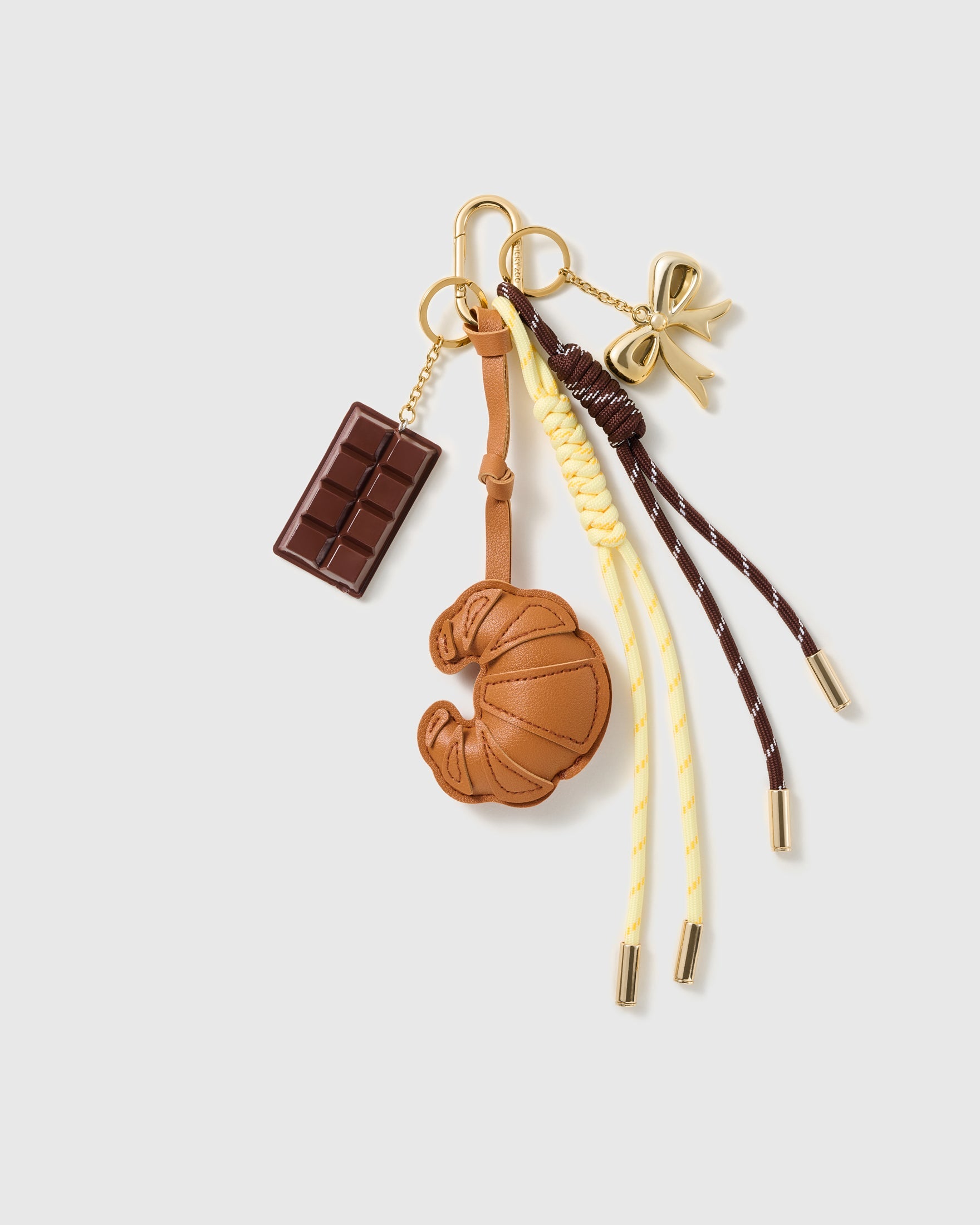 Croissant Chocolate Stack Charm with a croissant, chocolate bar, bow detail and yellow-and-brown cords on a light background