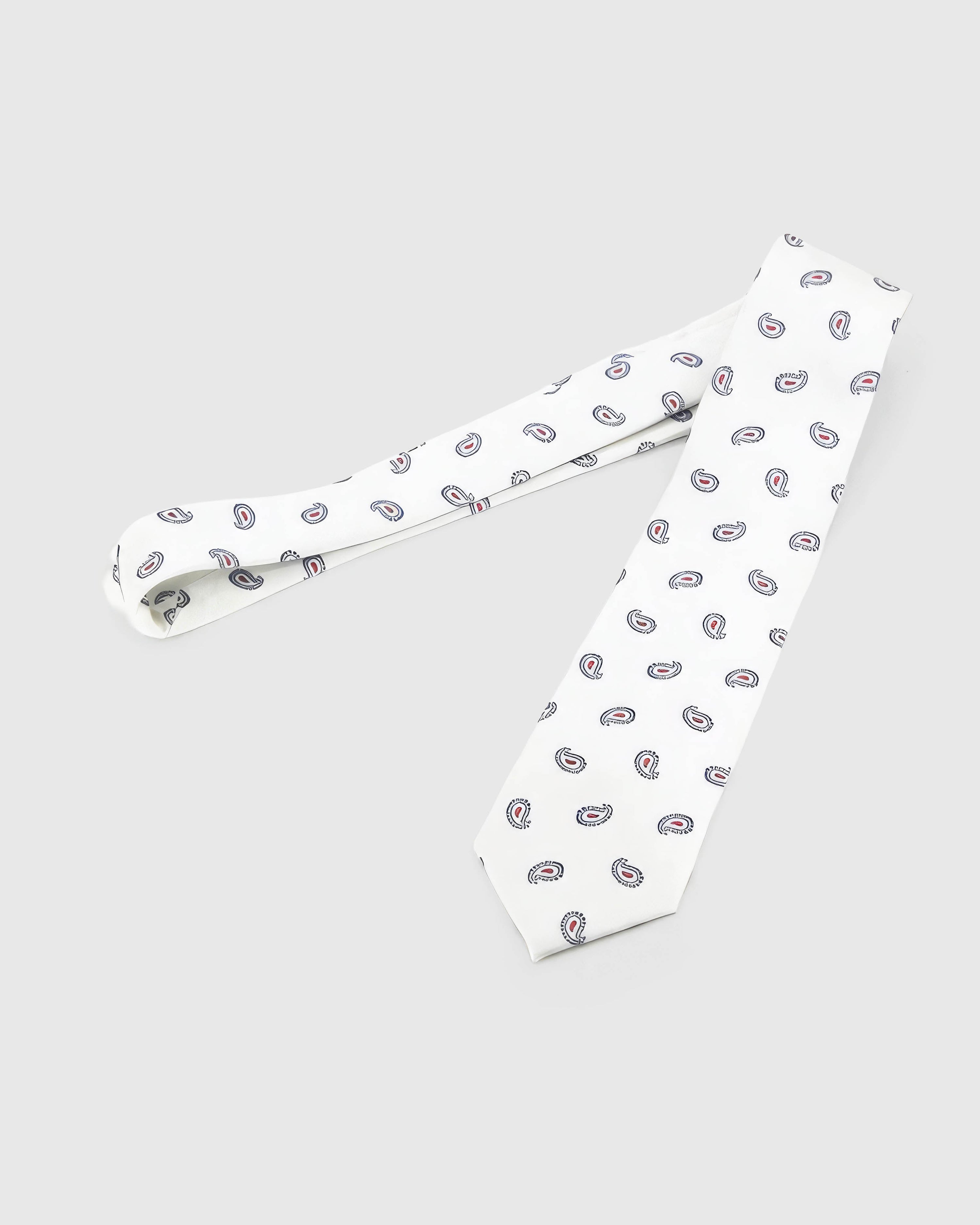 Ivory Tricolor Paisley Tie laid full length on white, showing an ivory ground with small navy-outline paisleys and crisp crimson centres from blade to tail.