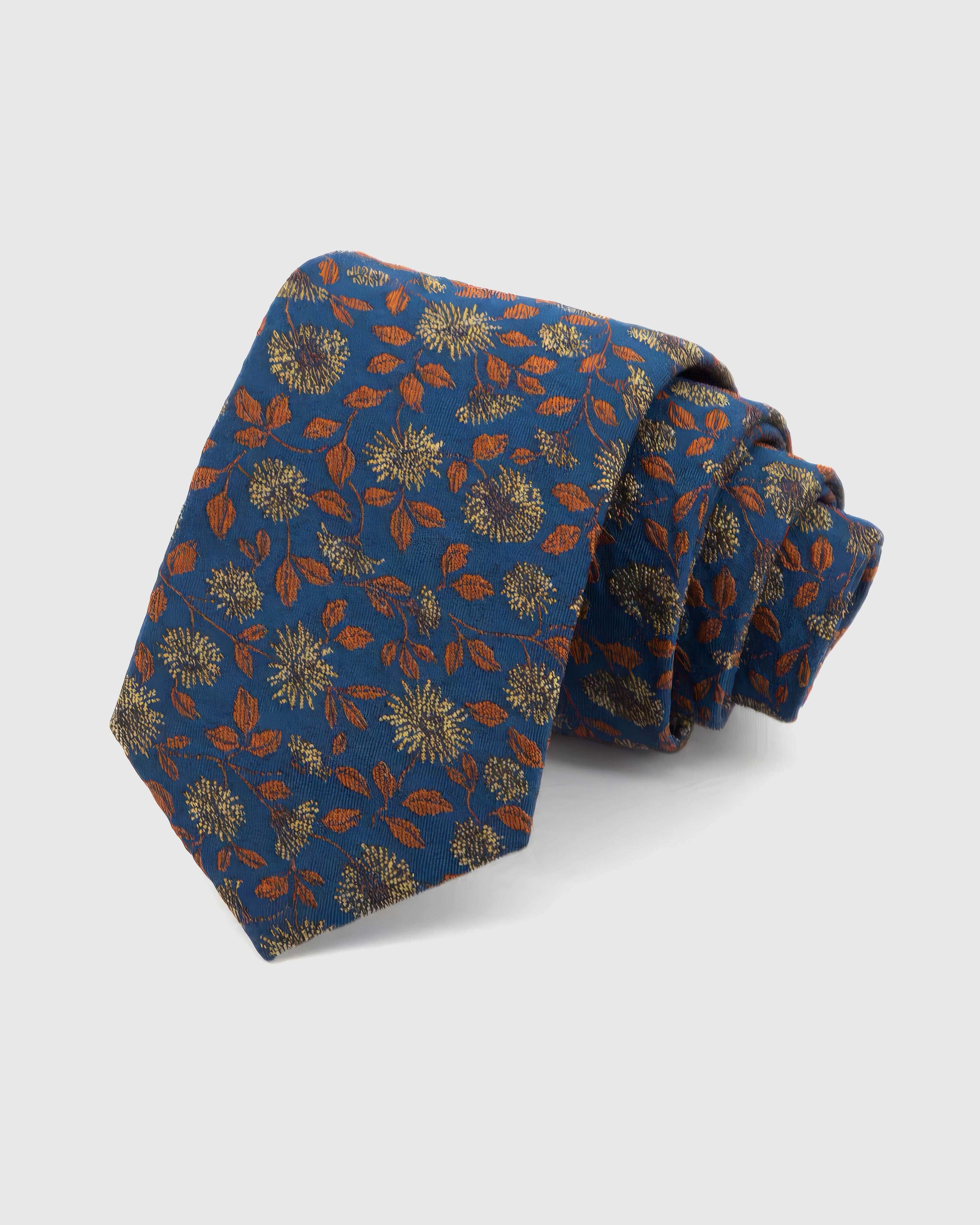 STICKYZOO Cobalt Copper Bloom Tie rolled on white, showing a cobalt ground with copper-toned leaves and pale-gold botanical blooms across the front blade.