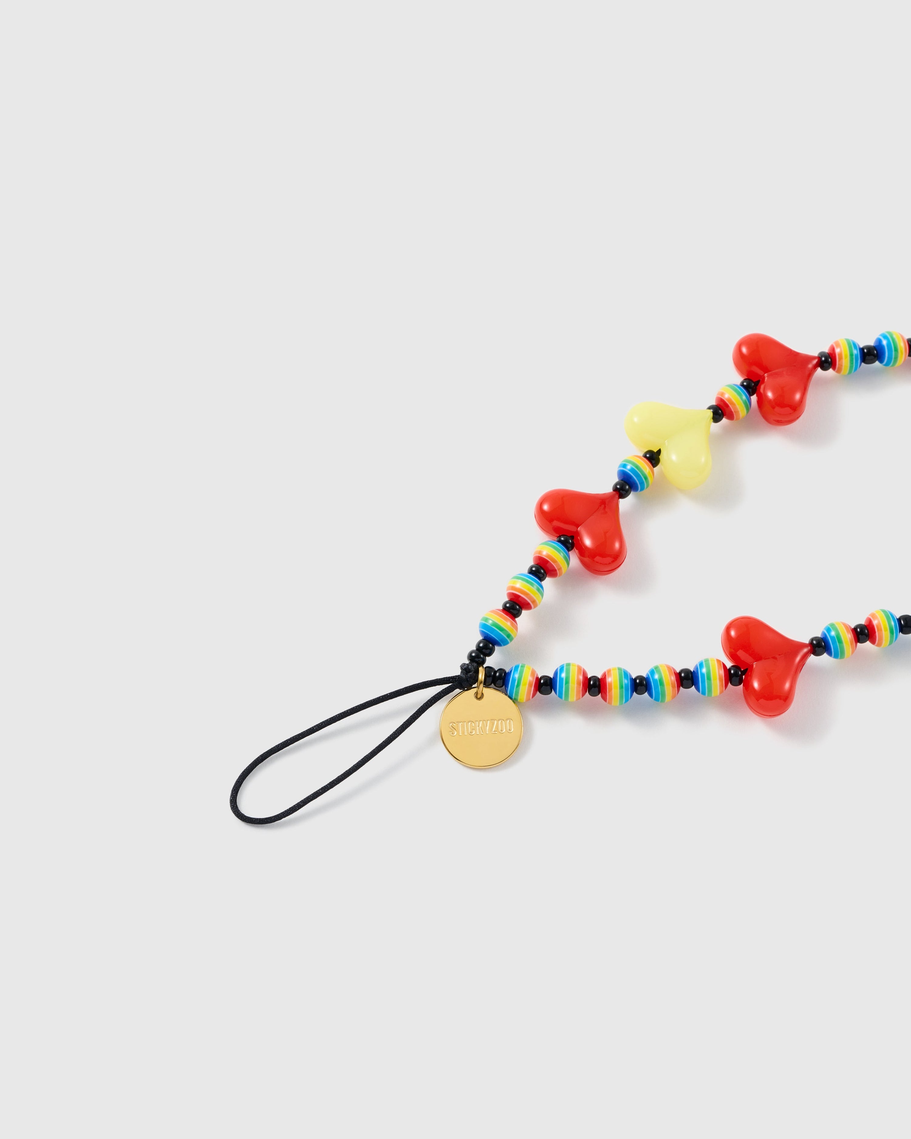 Close-up of the black stretch-cord loop and gold STICKYZOO disc beside glossy red and yellow hearts and rainbow-striped beads.