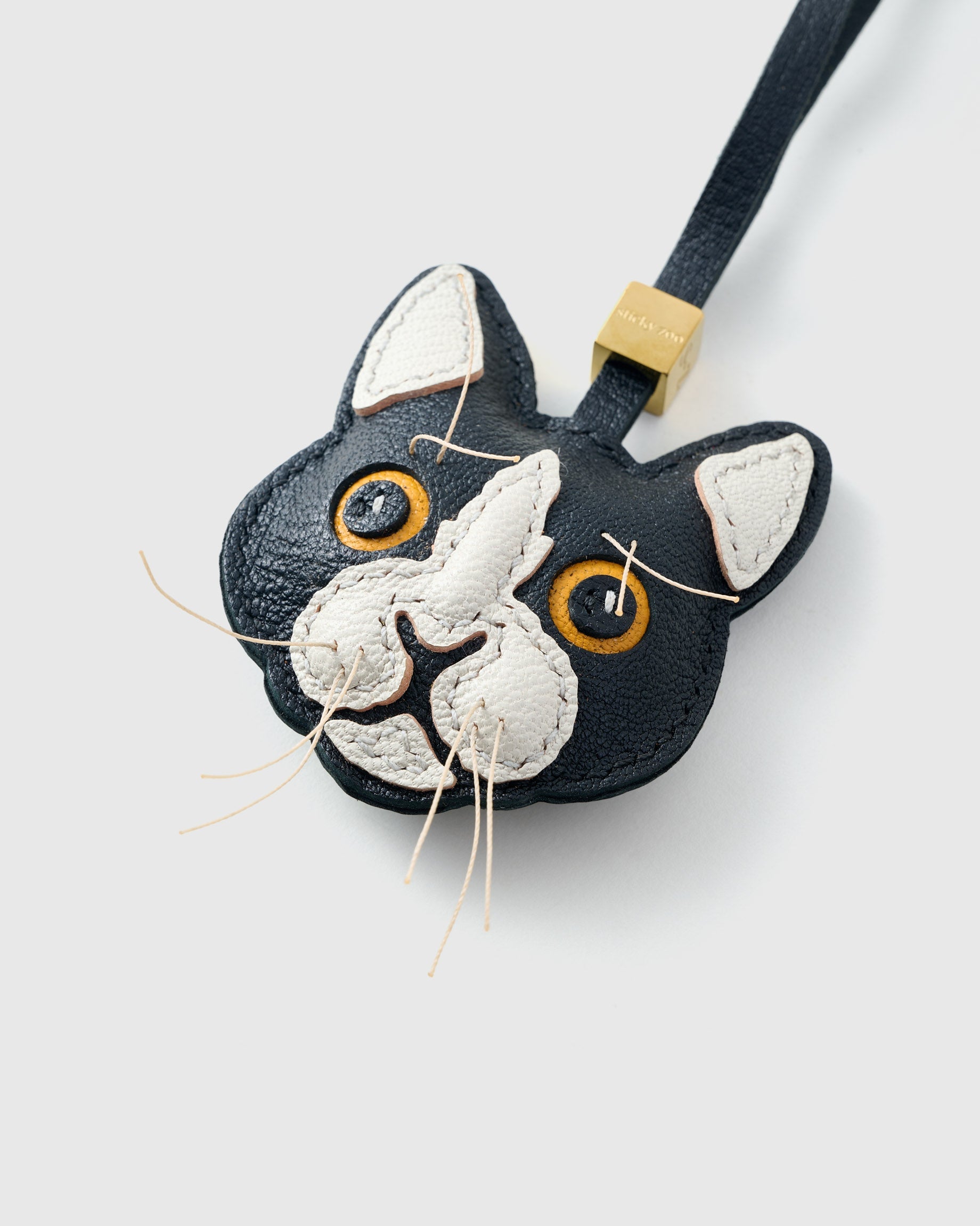 Tight close-up of the tuxedo cat face — gold-yellow eyes in real leather, white face mask, real fiber whiskers stitched in. Every panel by hand.