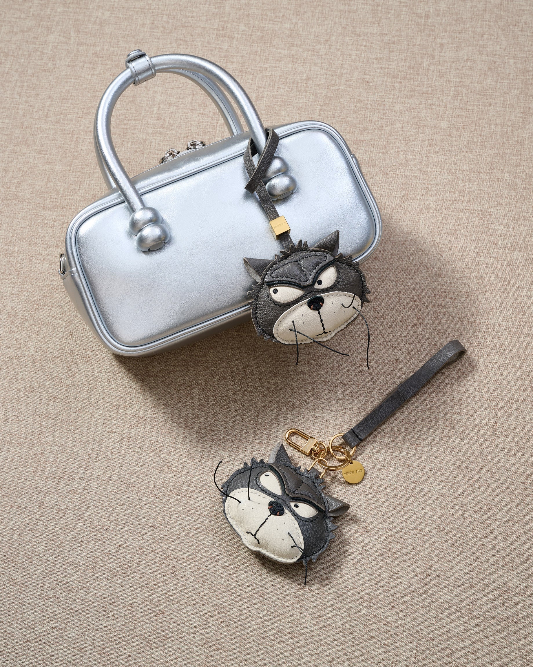 Overhead flatlay — Grump cat charm in strap and keychain editions, alongside a metallic silver box bag. Finished one at a time, by hand.
