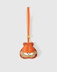 Garfield-style charm handmade from French Alran genuine goatskin — orange tabby face with darker stitched stripes, half-closed eyes, sandy muzzle with small frown, orange leather strap. Three days by hand. Arrives in luxury gift packaging. Designed in Amsterdam.