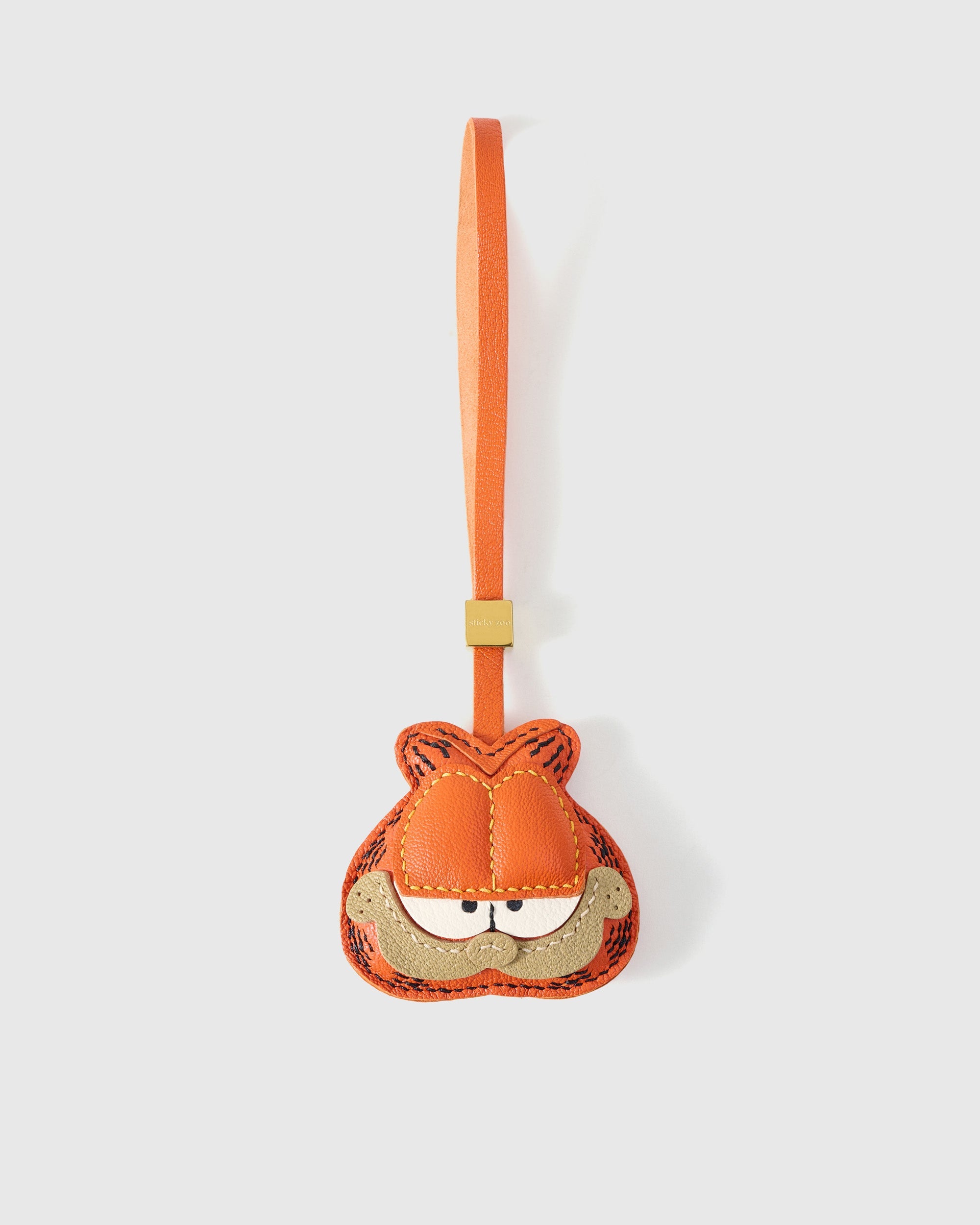 Garfield-style charm handmade from French Alran genuine goatskin — orange tabby face with darker stitched stripes, half-closed eyes, sandy muzzle with small frown, orange leather strap. Three days by hand. Arrives in luxury gift packaging. Designed in Amsterdam.