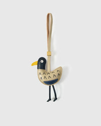 Gull charm handmade from French Alran genuine goatskin — cream-bronze body, black head, yellow beak, navy chest accent, black dangling legs, tan leather strap. Three days by hand. Arrives in luxury gift packaging. Designed in Amsterdam.
