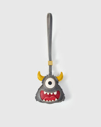 Little monster charm handmade from French Alran genuine goatskin — gray body, single large white eye, yellow horns, wide red mouth with hand-cut white teeth, gray leather strap. Three days by hand. Arrives in luxury gift packaging. Designed in Amsterdam.