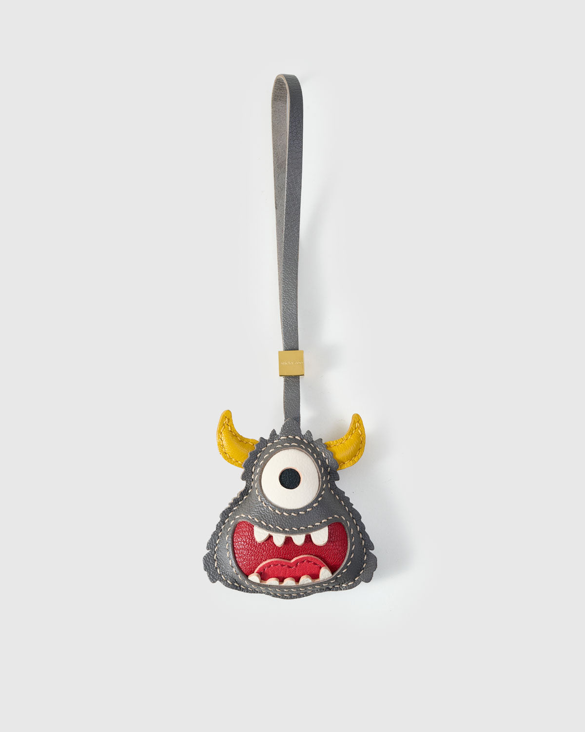 Little monster charm handmade from French Alran genuine goatskin — gray body, single large white eye, yellow horns, wide red mouth with hand-cut white teeth, gray leather strap. Three days by hand. Arrives in luxury gift packaging. Designed in Amsterdam.