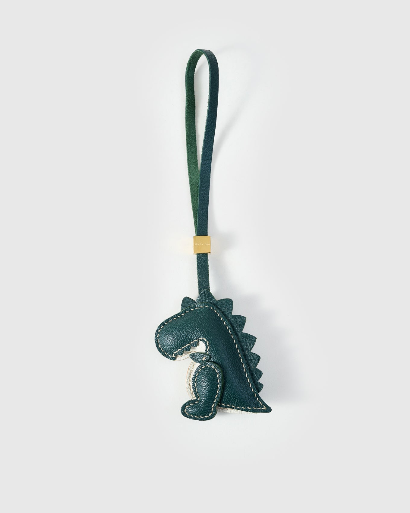 Green dinosaur charm handmade from French Alran genuine goatskin — deep forest-green body, cream belly, hand-cut spine ridges along the back, green leather strap. Three days by hand. Designed in Amsterdam.