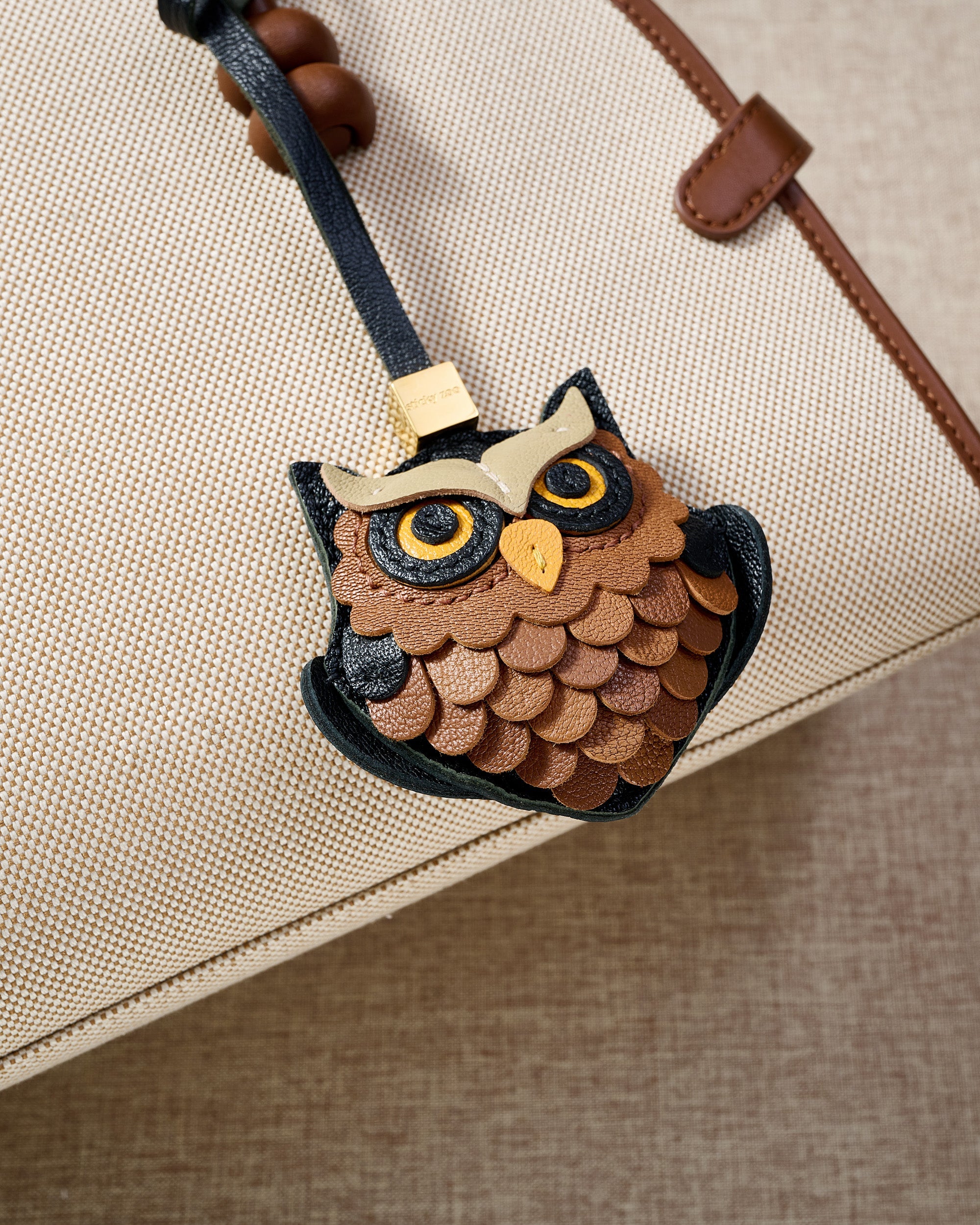 Close-up of the owl charm on a cream canvas bag — every scalloped feather layer cut and hand-stitched in real French leather, yellow eyes in real leather, yellow beak.