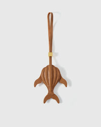 Sculpted caramel whale charm handmade from French Alran genuine goatskin — dimensional 3D body viewed from above, side fins, split tail fluke, cream hand-stitched seams, tan leather strap. Three days by hand. Arrives in luxury gift packaging. Designed in Amsterdam.