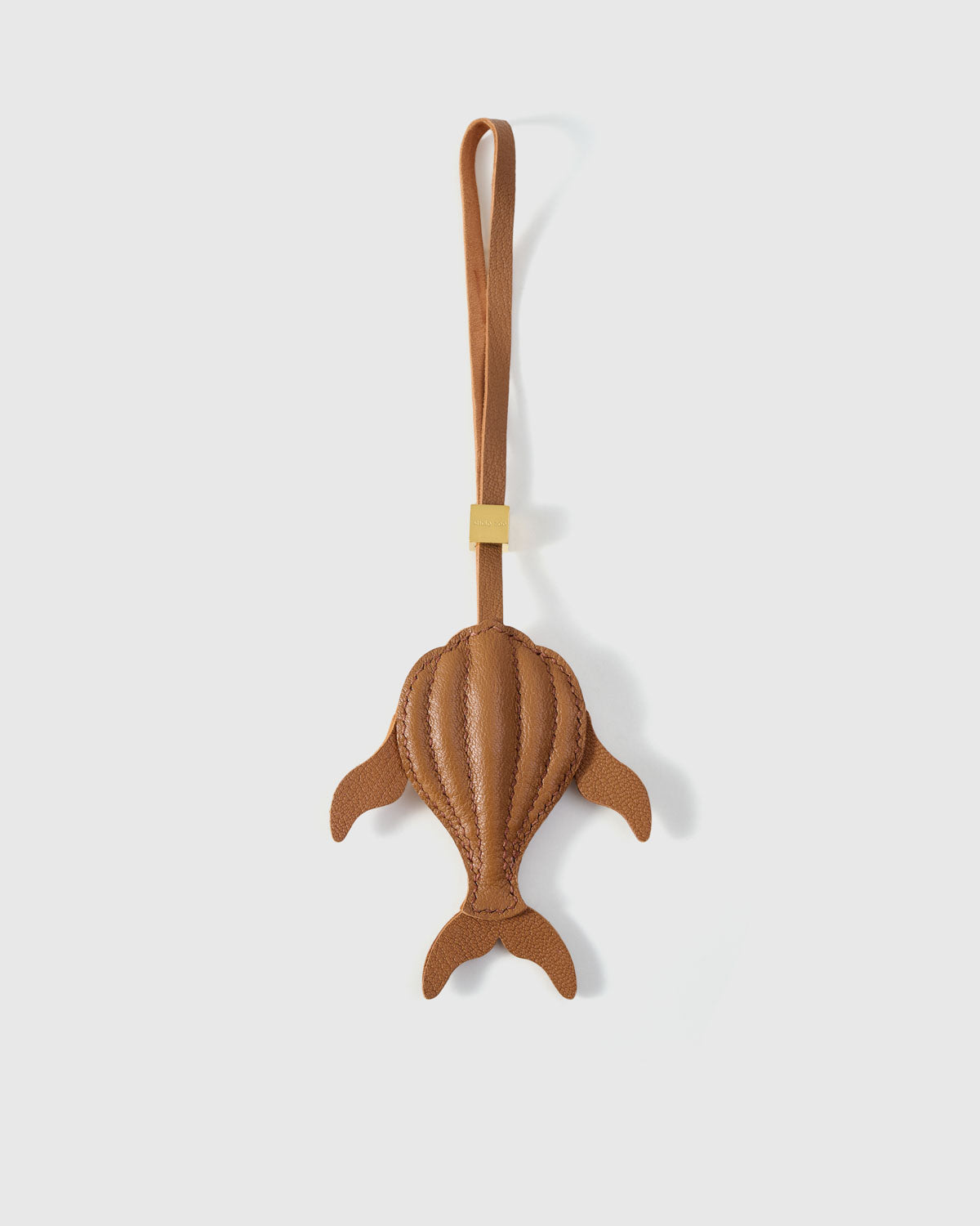 Sculpted caramel whale charm handmade from French Alran genuine goatskin — dimensional 3D body viewed from above, side fins, split tail fluke, cream hand-stitched seams, tan leather strap. Three days by hand. Arrives in luxury gift packaging. Designed in Amsterdam.