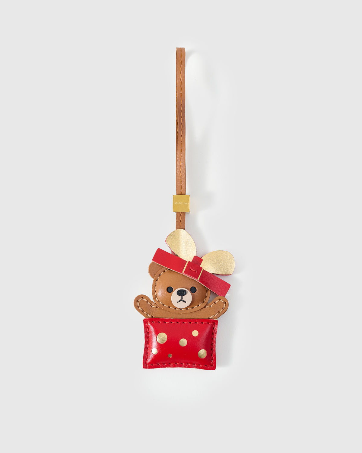 Teddy Bear Gift charm handmade from French Alran genuine goatskin — caramel teddy bear peeking out of a red gift box with gold polka dots, golden metallic bow, red leather ribbon, paws gripping the edge, tan leather strap. Three days by hand. Arrives in luxury gift packaging. Designed in Amsterdam.