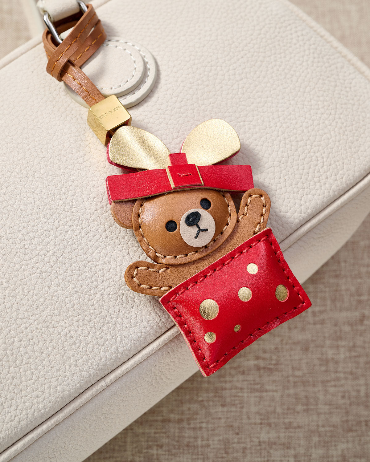 Teddy Bear Gift charm close on a cream pebbled-leather bag — a bear peeking out of a red-and-gold box, made from real French leather. The gift that says something before they even unwrap it.