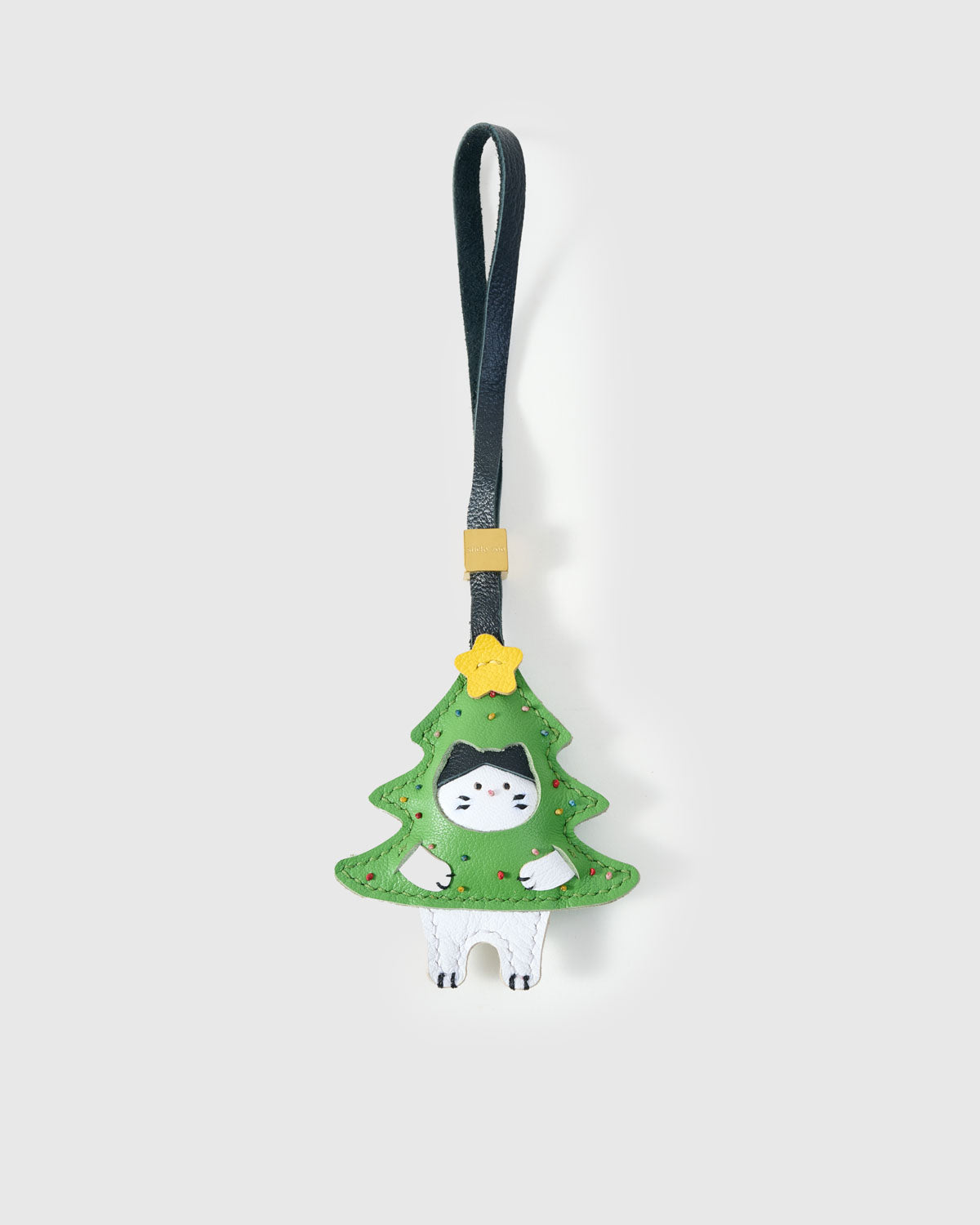 Kitten in Christmas Tree charm handmade from French Alran genuine goatskin — green tree silhouette with a black-and-white kitten peeking out, hand-stitched ornaments, gold star, black leather strap. Three days by hand. Arrives in luxury gift packaging. Designed in Amsterdam.