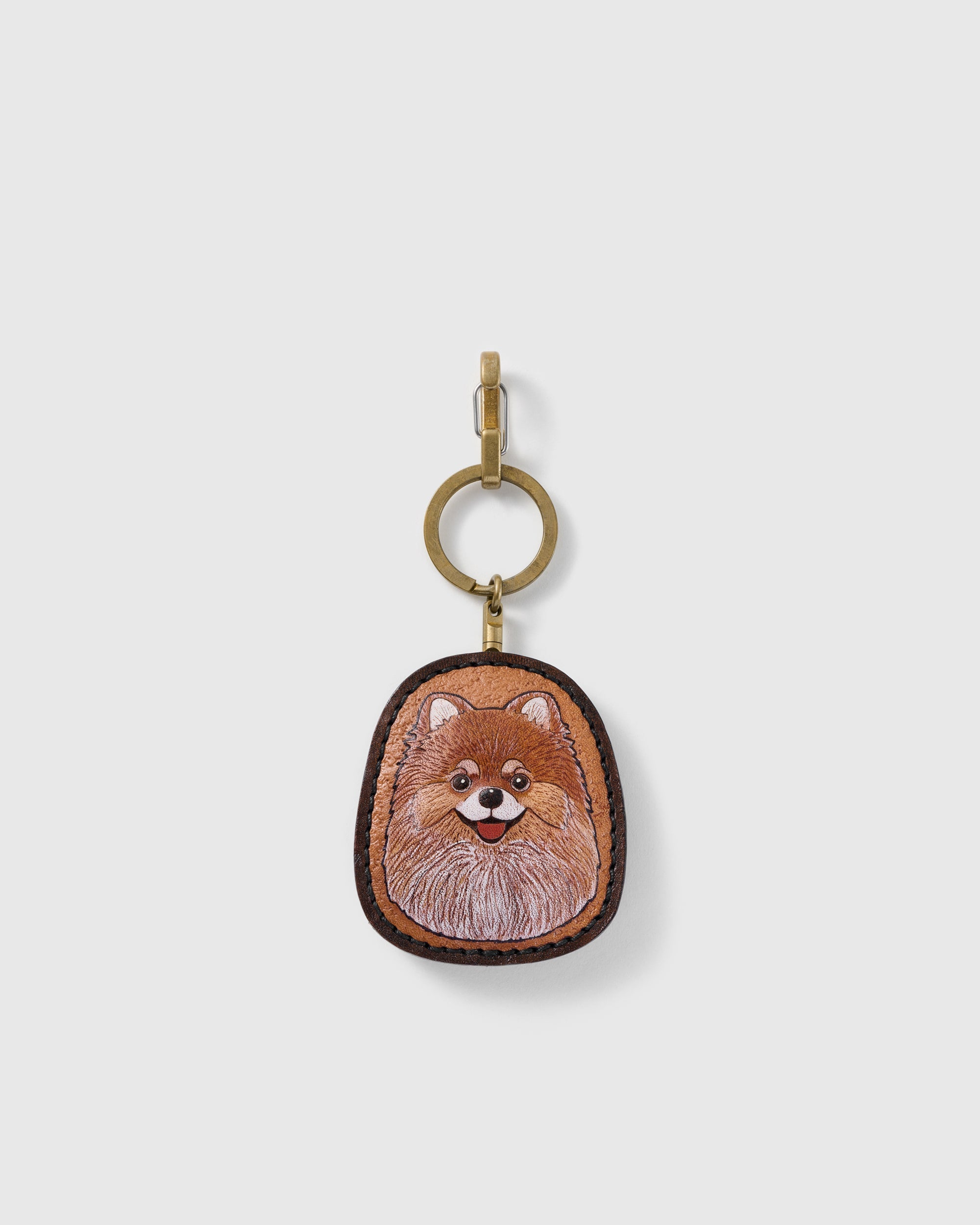 STICKYZOO Posie Pomeranian portrait charm with a smiling warm tan and cream face, dark stitched border, and gold-tone ring-and-clip hardware on a pale background
