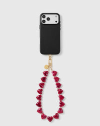 Black phone case with the ruby-red heart bead wrist strap looping below, on an 18K gold-plated hook and engraved disc.