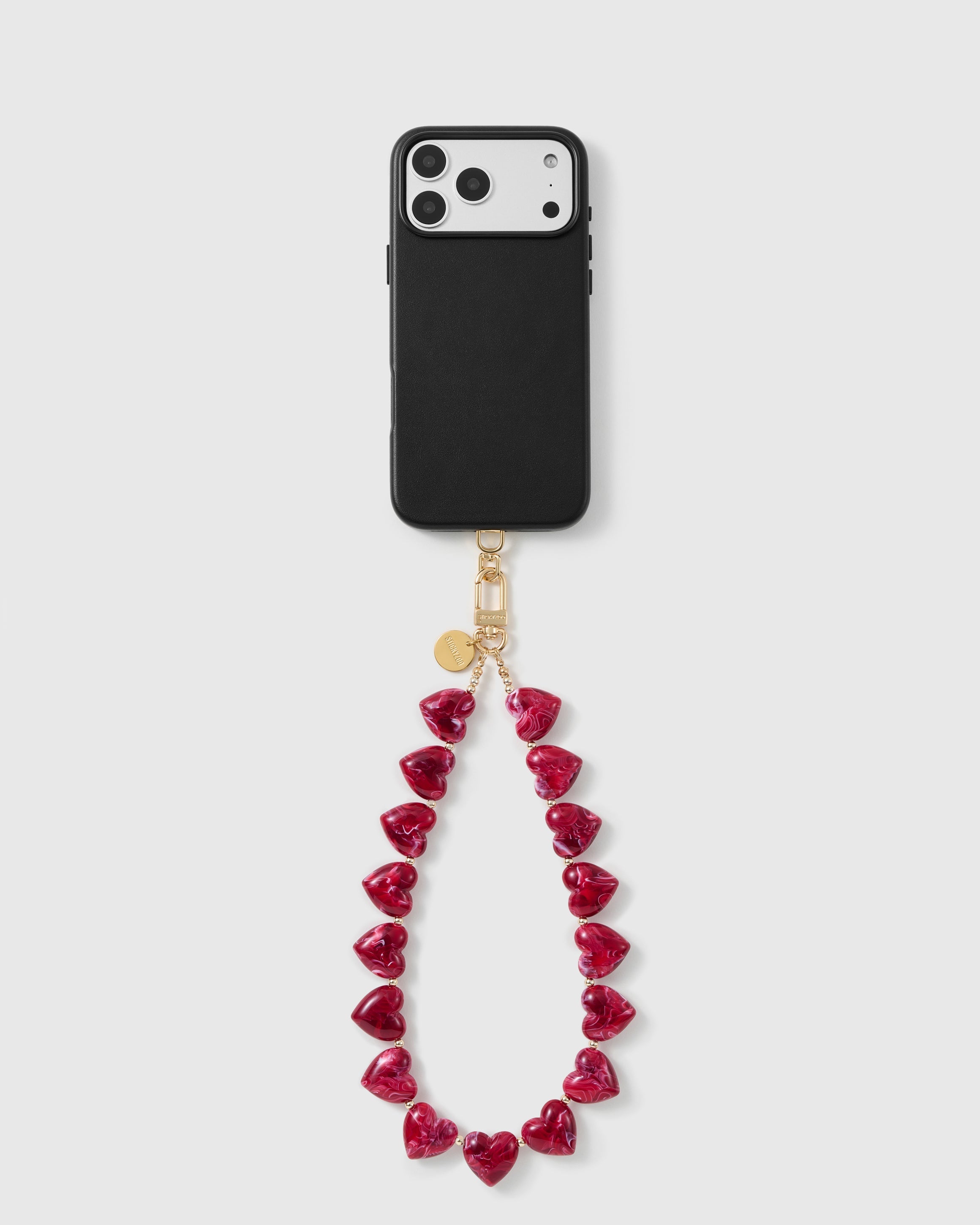 Black phone case with the ruby-red heart bead wrist strap looping below, on an 18K gold-plated hook and engraved disc.