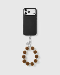 Hazelnut velvet pom wrist strap clipped to a black phone case for hands-free carry