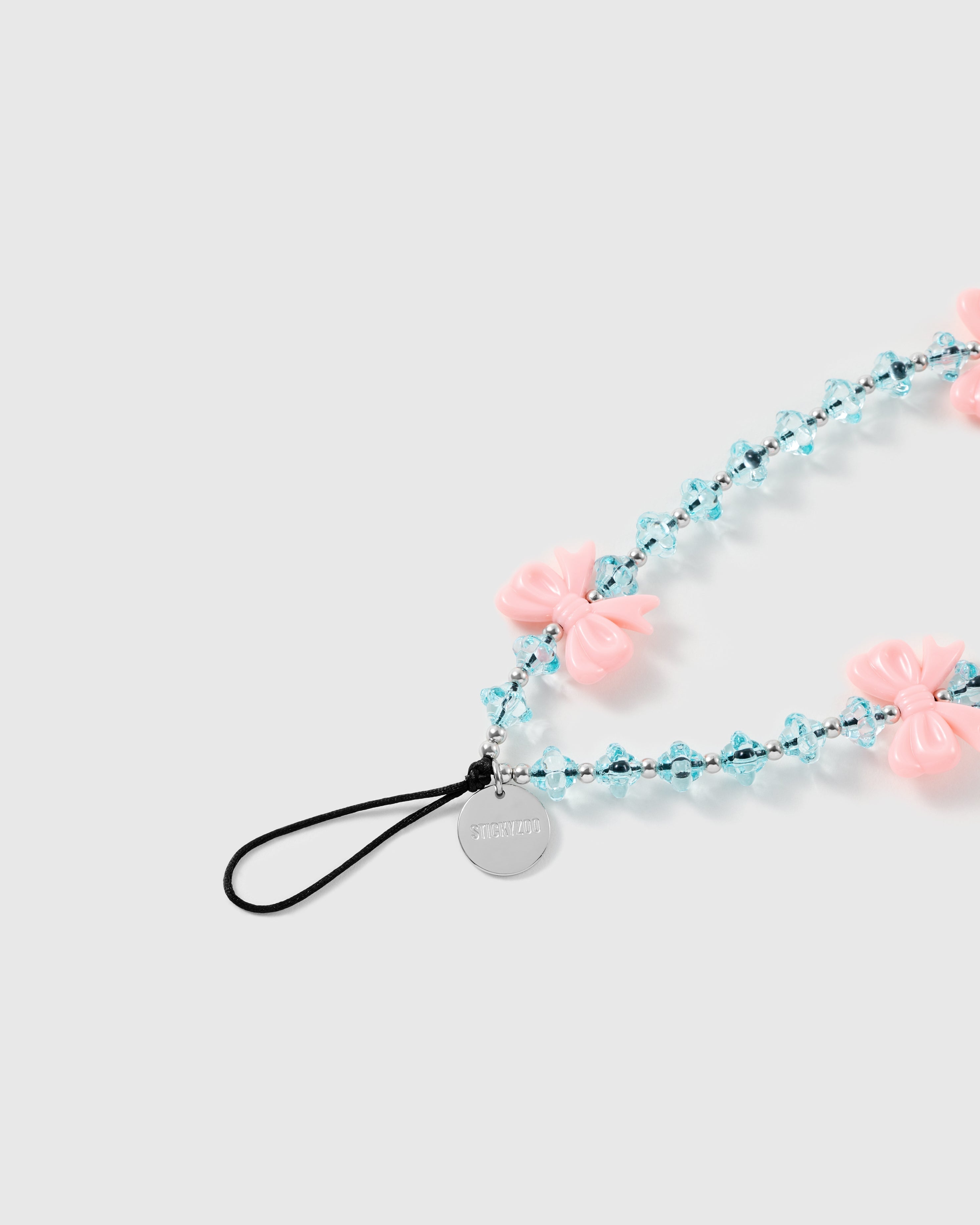 Close-up of the STICKYZOO strap's black stretch-cord loop and engraved silver-tone disc beside glossy pink bows and faceted ice-blue beads