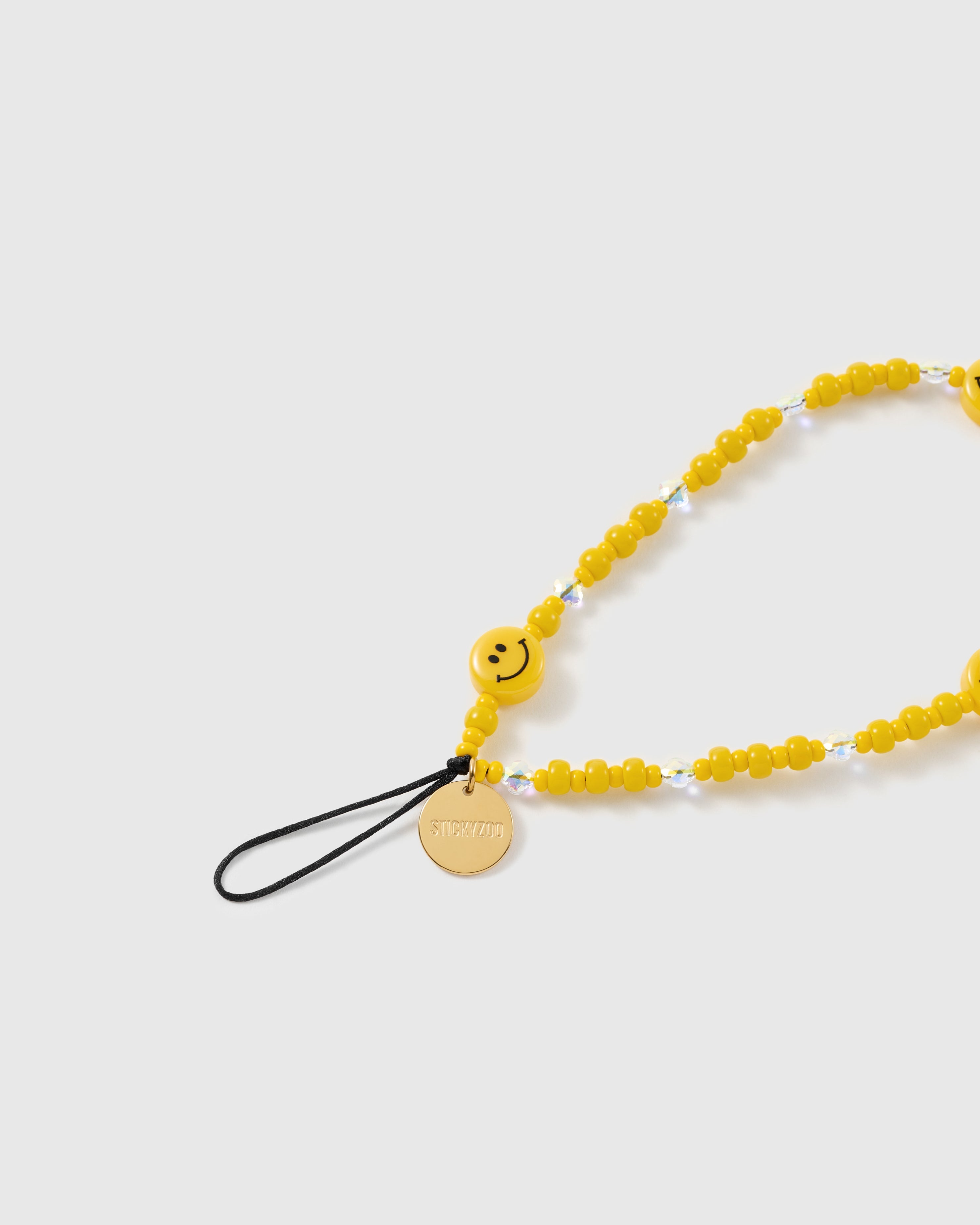 Close-up of the gold-tone STICKYZOO disc on a black stretch-cord loop, beside a yellow smiley bead and faceted clear accent beads.