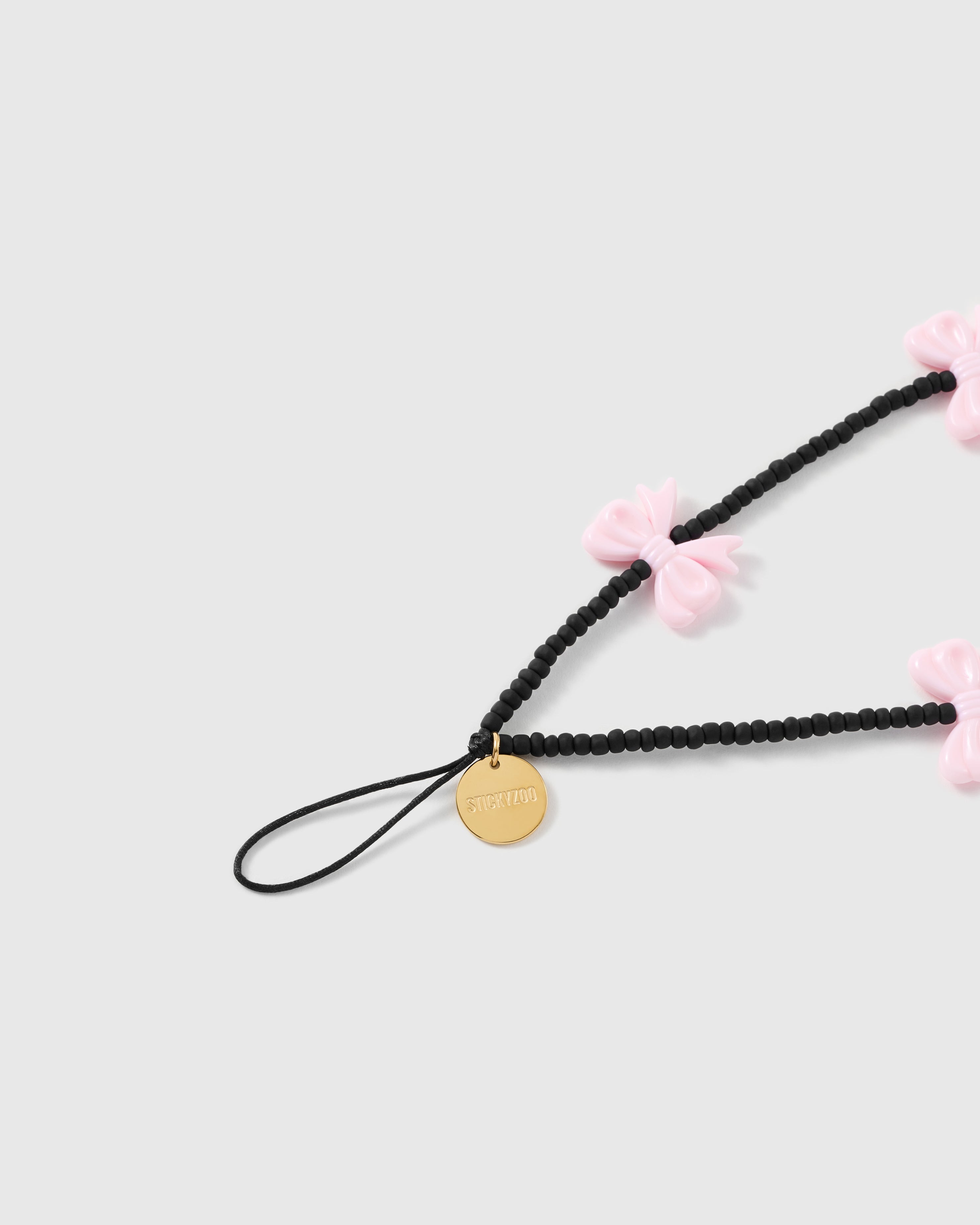 Close-up of the gold-tone STICKYZOO disc hanging where the black stretch-cord loop meets the matte black beads, with glossy pink bows spaced along the strands.