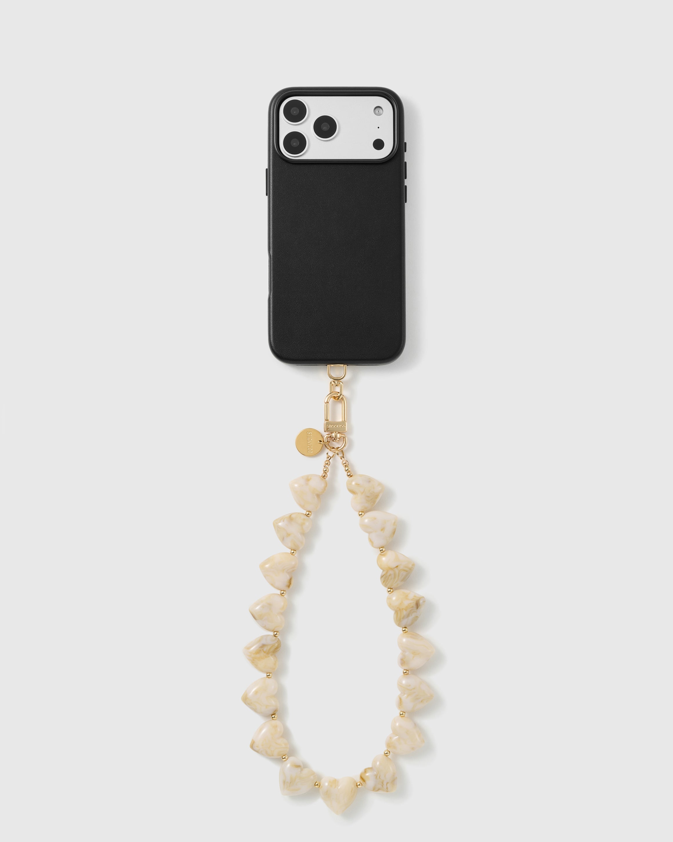 Black triple-camera phone case with the ivory heart bead wrist strap hanging in a loop from a gold hook and engraved gold disc.