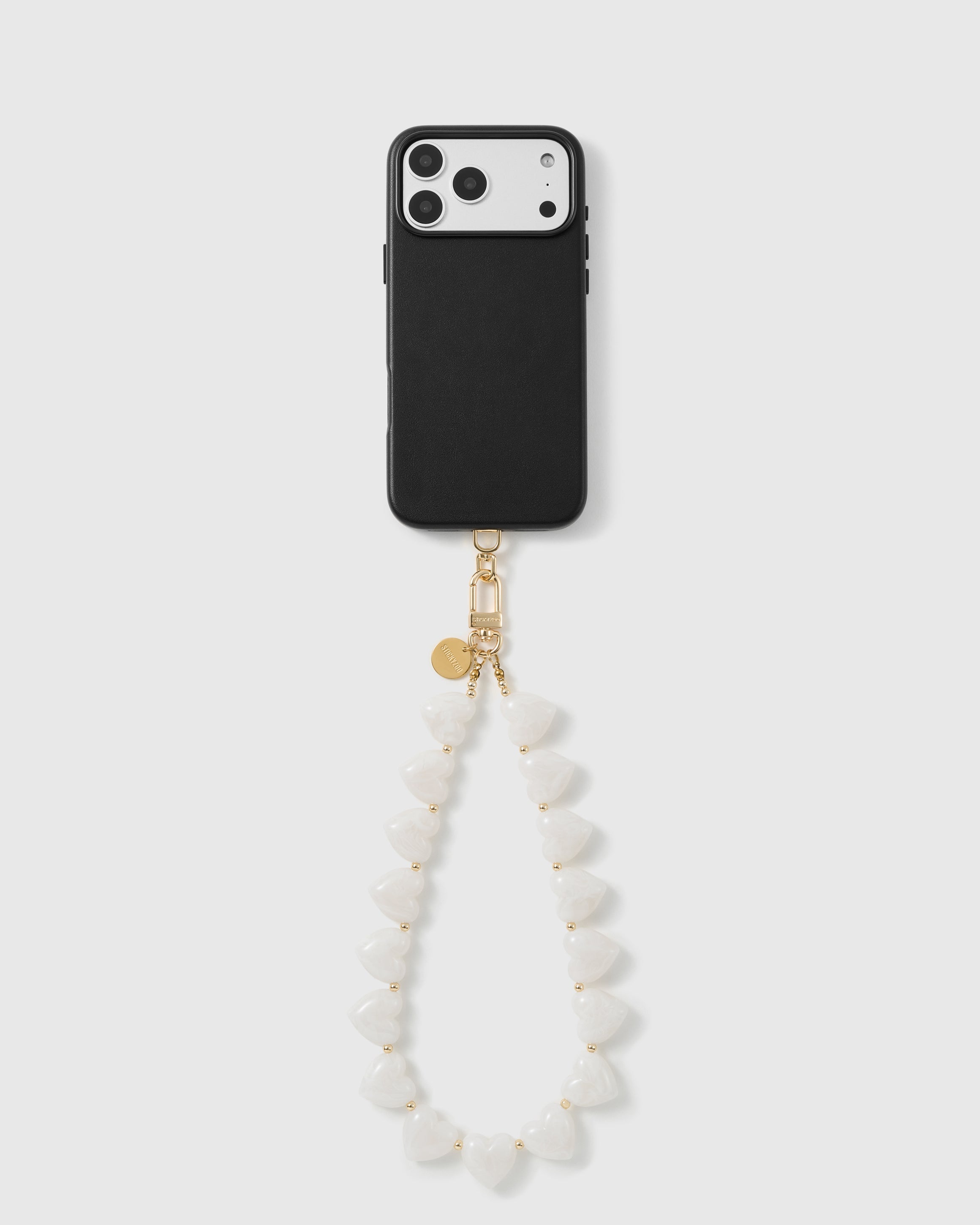 Black phone with the milky white heart bead wrist strap hanging in a soft loop, gold hook and disc at the top.
