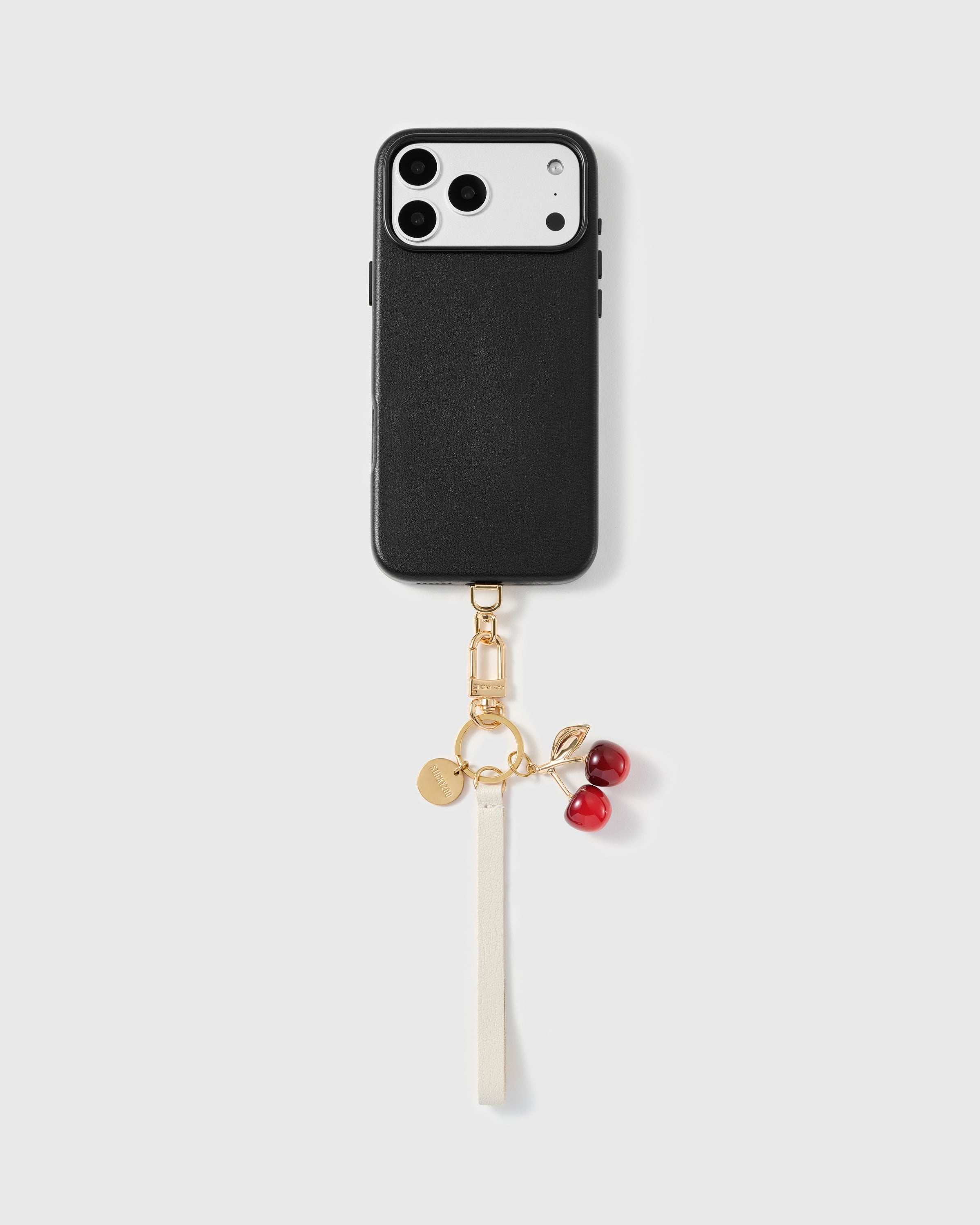 Ivory leather cherry phone strap clipped to a black leather phone case, the glossy cherry charm and cream tail hanging at the base.