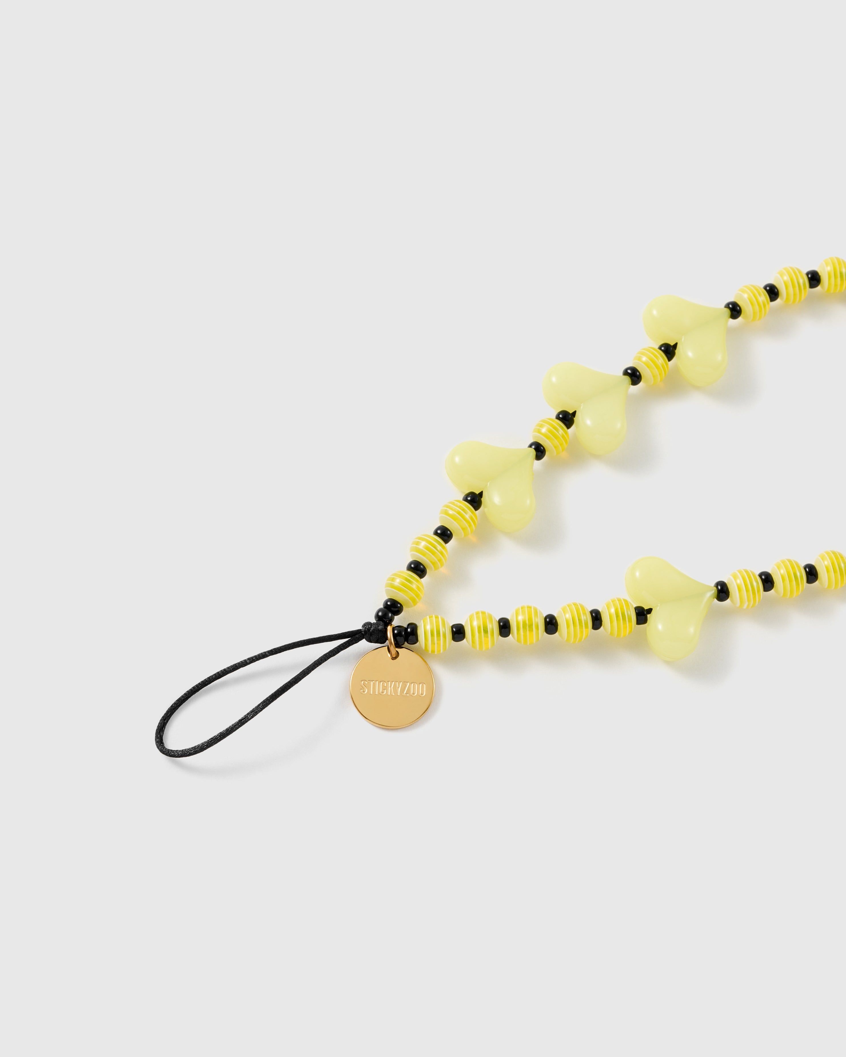 Close-up of the gold-tone STICKYZOO disc on the black stretch-cord loop, beside striped yellow beads and a pale lemon heart bead