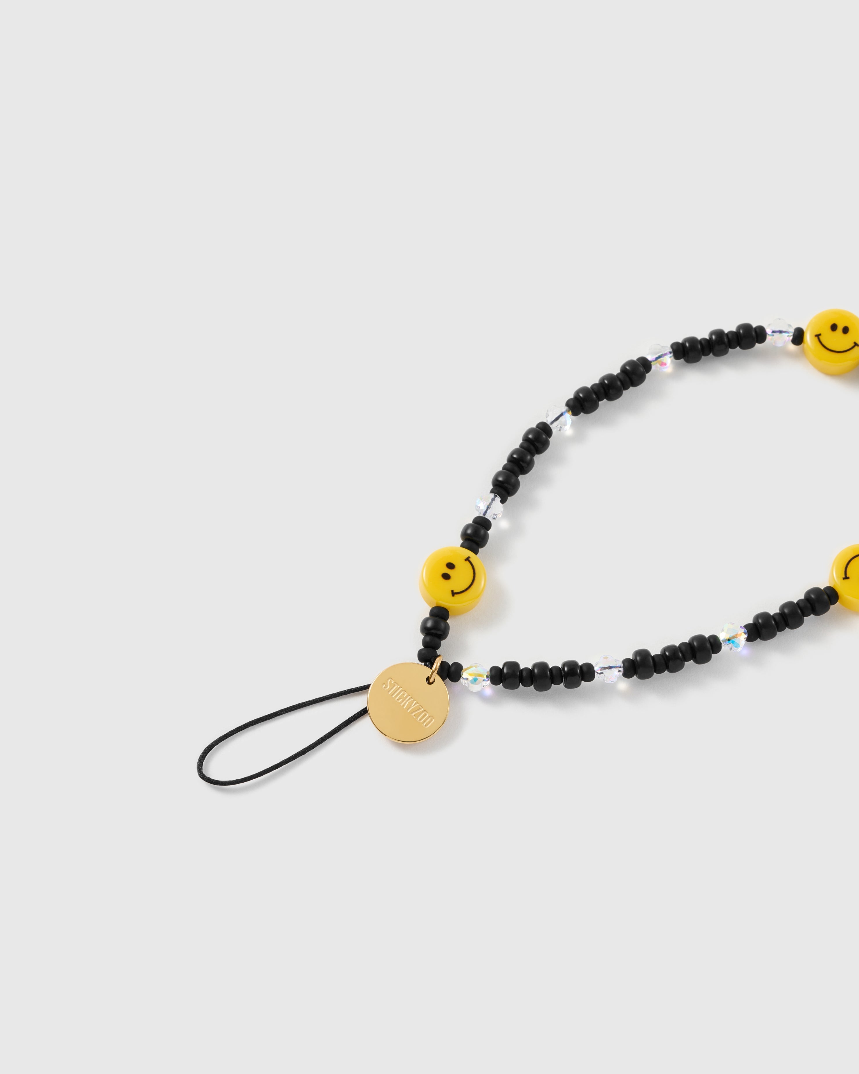 Close-up of the STICKYZOO gold-tone disc and black stretch-cord loop beside a yellow smiley bead and faceted clear beads with an iridescent shimmer.