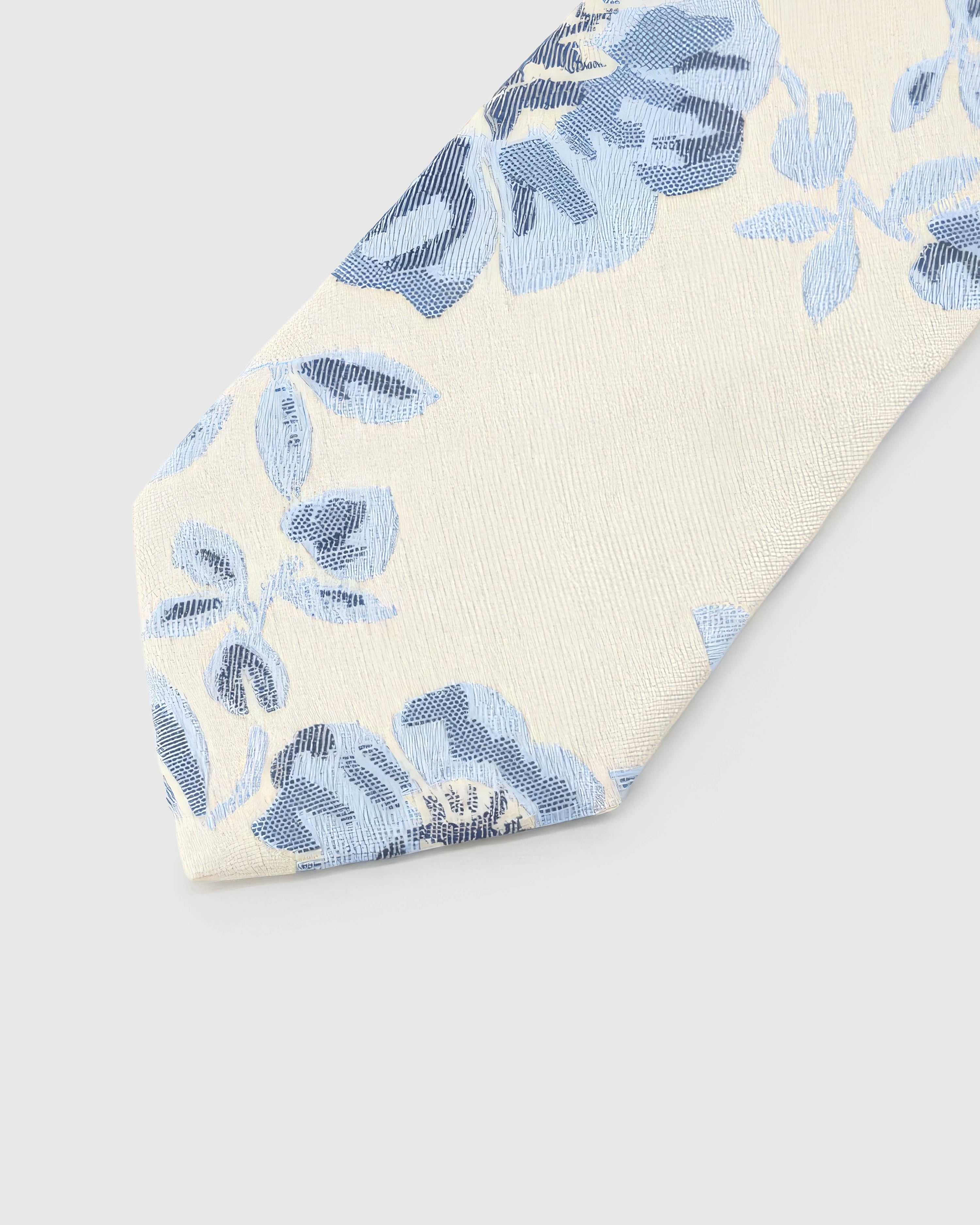 Ivory Porcelain Bloom Tie detail showing powder-blue and indigo botanical blooms, raised thread and the tactile ivory silk surface.
