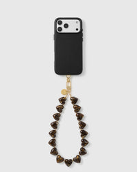 Tortoiseshell Heart wrist strap looped below a black phone case, with marbled amber heart beads, a gold-tone hook and engraved disc.