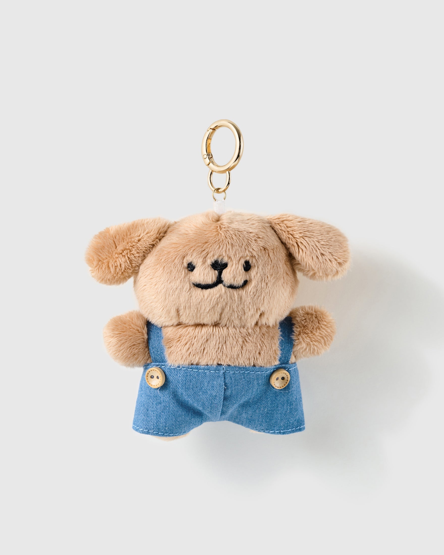 Denim Day Pup Charm, tan plush pup with blue denim-look overalls and button details, on a light background