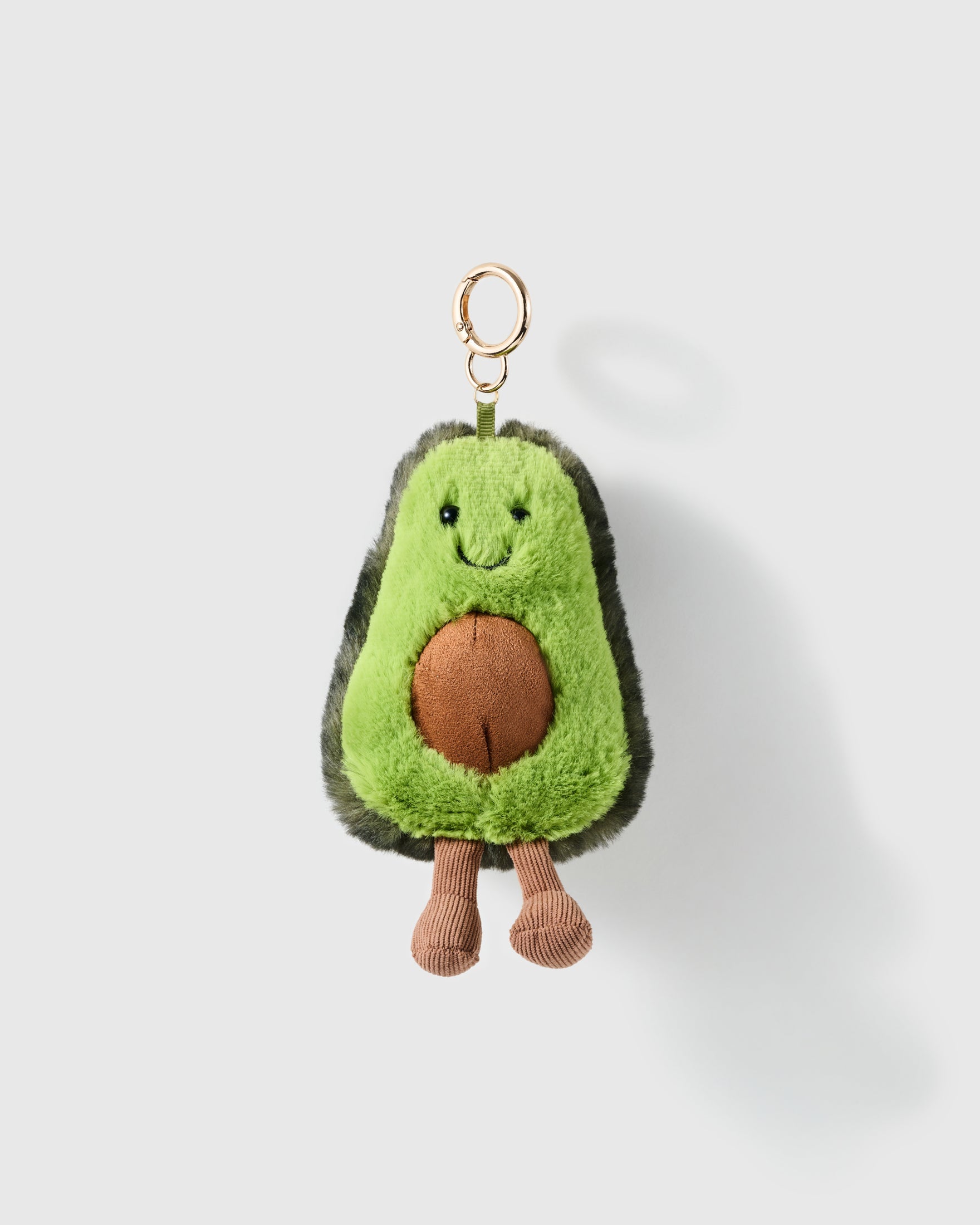 Happy Avo Charm, green plush avocado with brown centre, embroidered face and dangling legs, on a light background