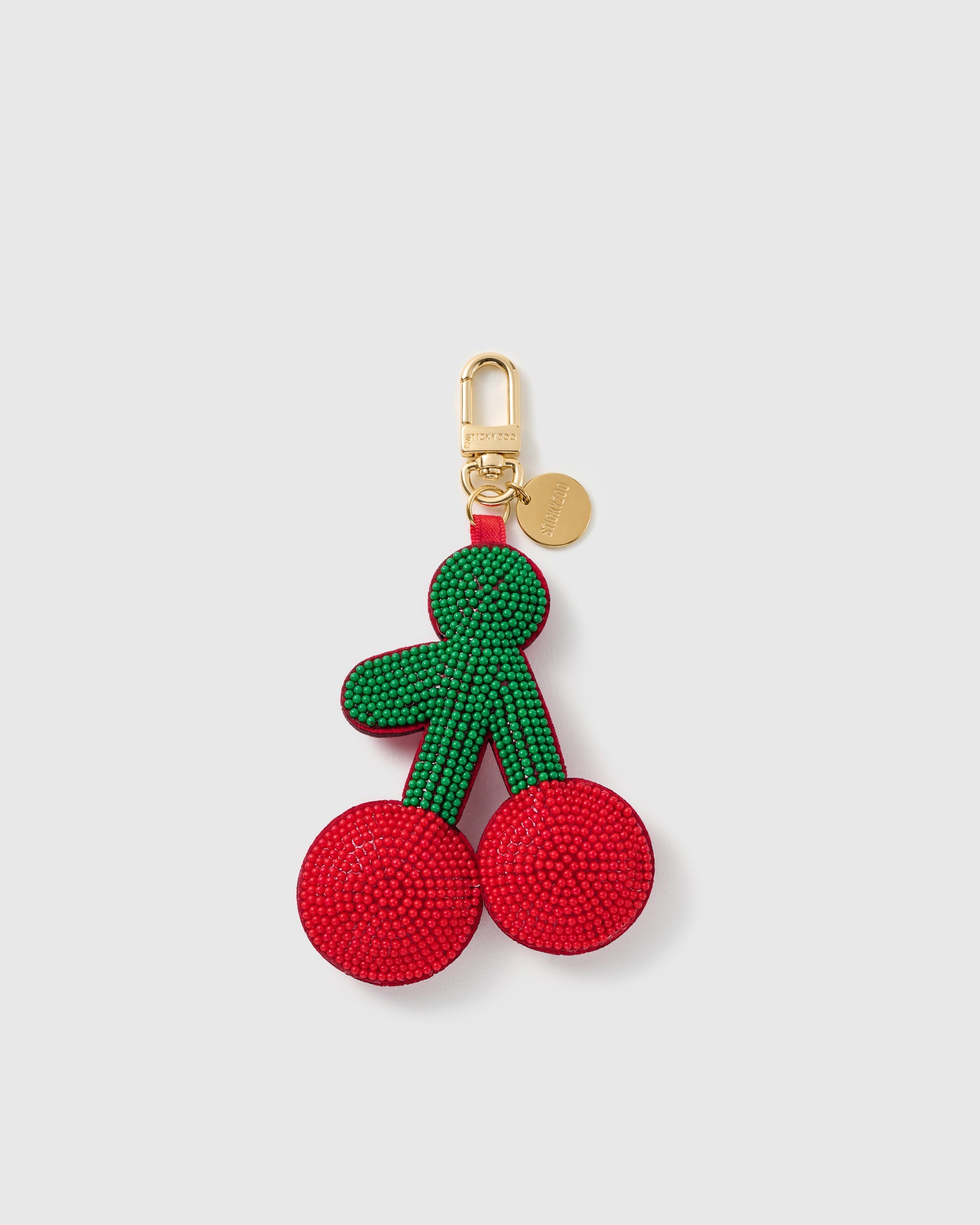 STICKYZOO Romy Cherry Duet charm formed as one joined piece, with two red bead-covered cherries, green stems and leaves, a gold-tone swivel clasp, and engraved tag on a pale background