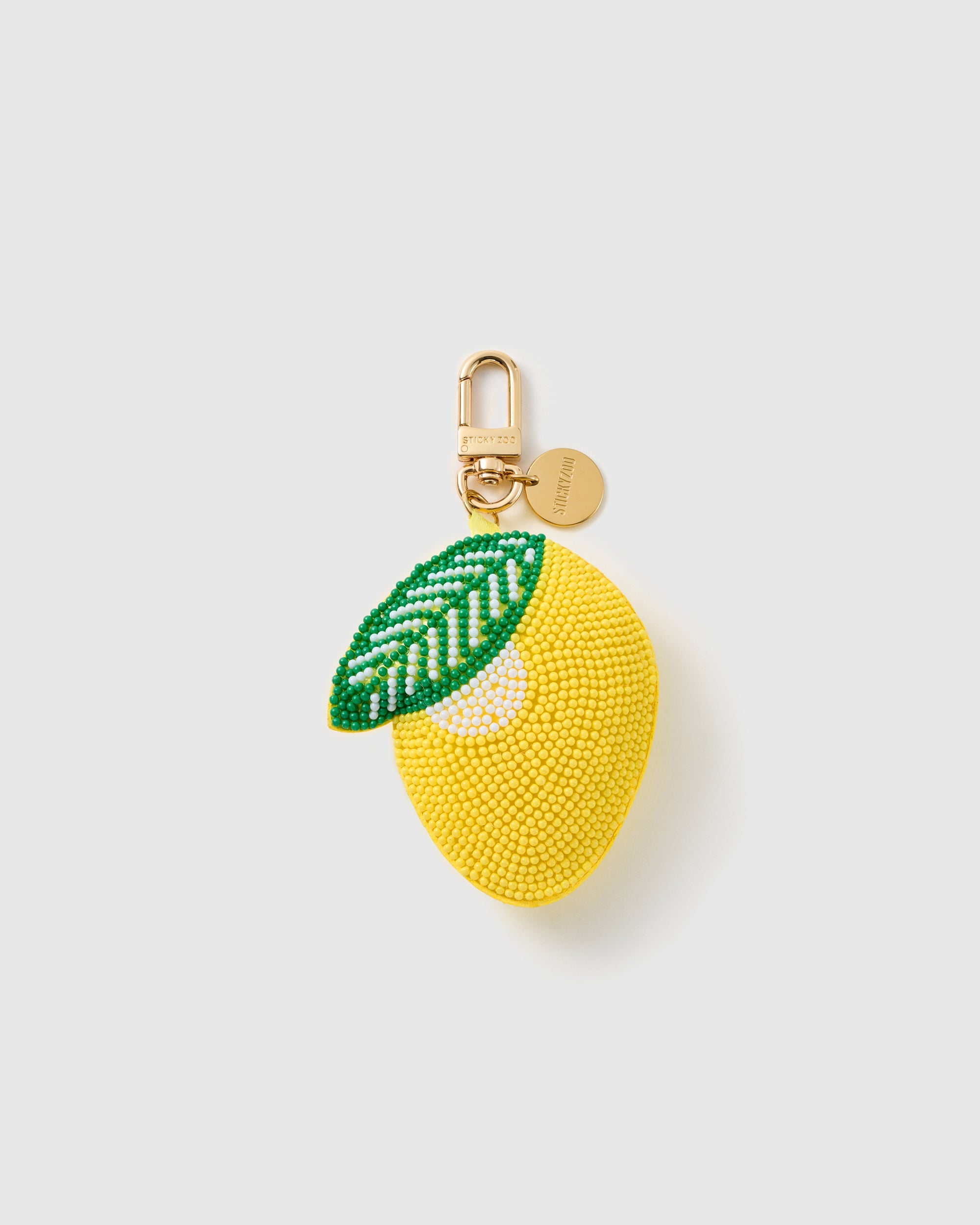 STICKYZOO Gigi Lemon Drop charm with a rounded yellow bead-covered fruit, green leaf, white accent pattern, gold-tone swivel clasp, and engraved round tag on a pale background