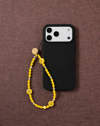 Yellow smiley bead phone strap looped onto a black phone case, its glossy beads and smiley faces bright against dark textured fabric.